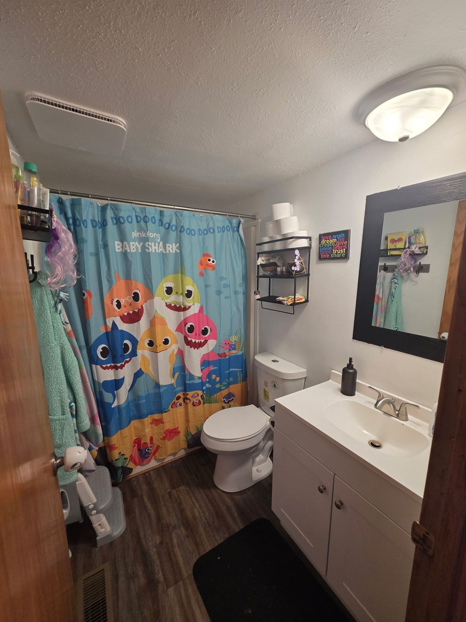 Bathroom with Baby Shark shower curtain, white vanity, and toilet. Dark flooring and black rug.