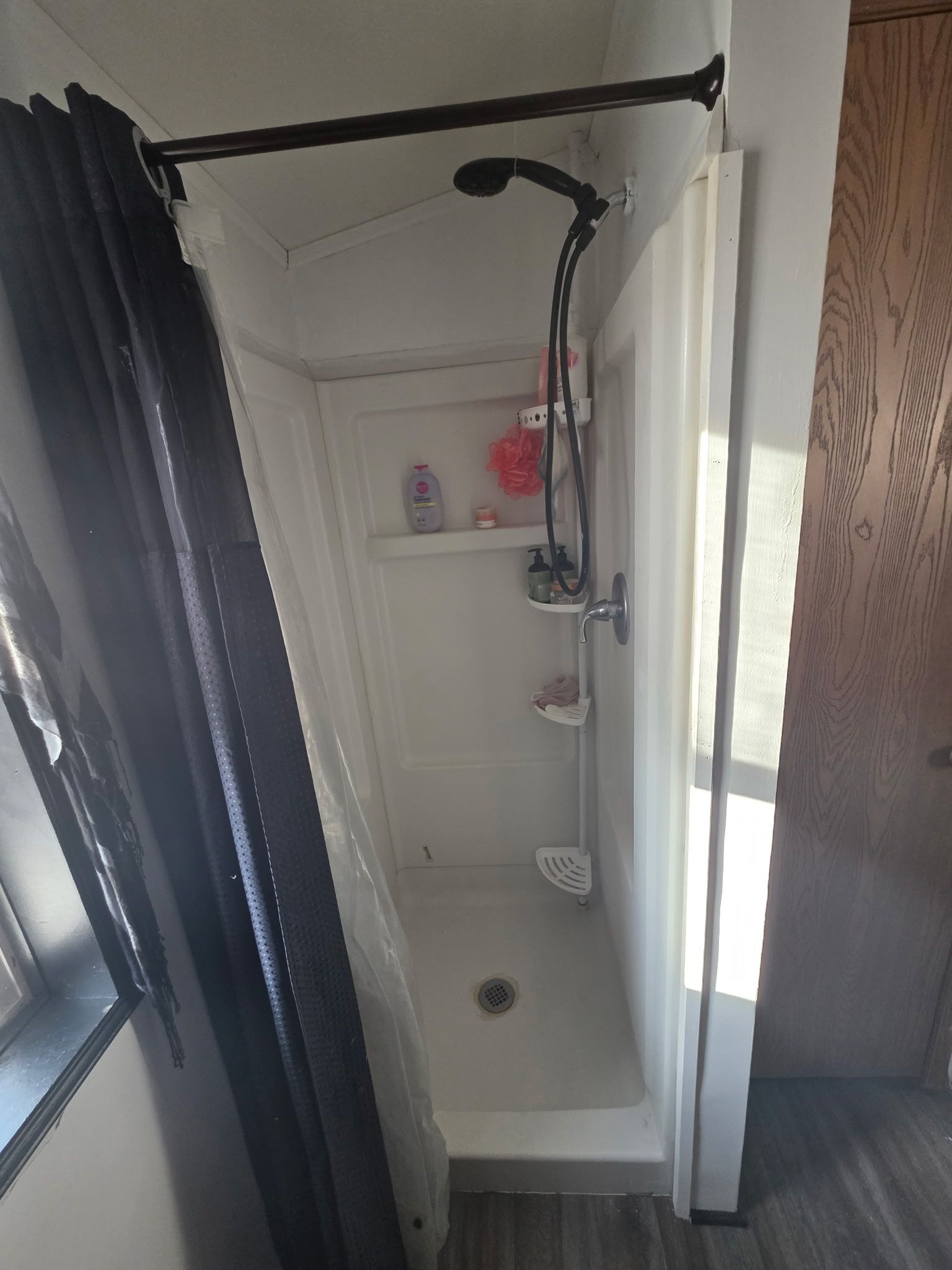 Small, white shower stall with a black showerhead and curtain rod. Dark shower curtain to the left.
