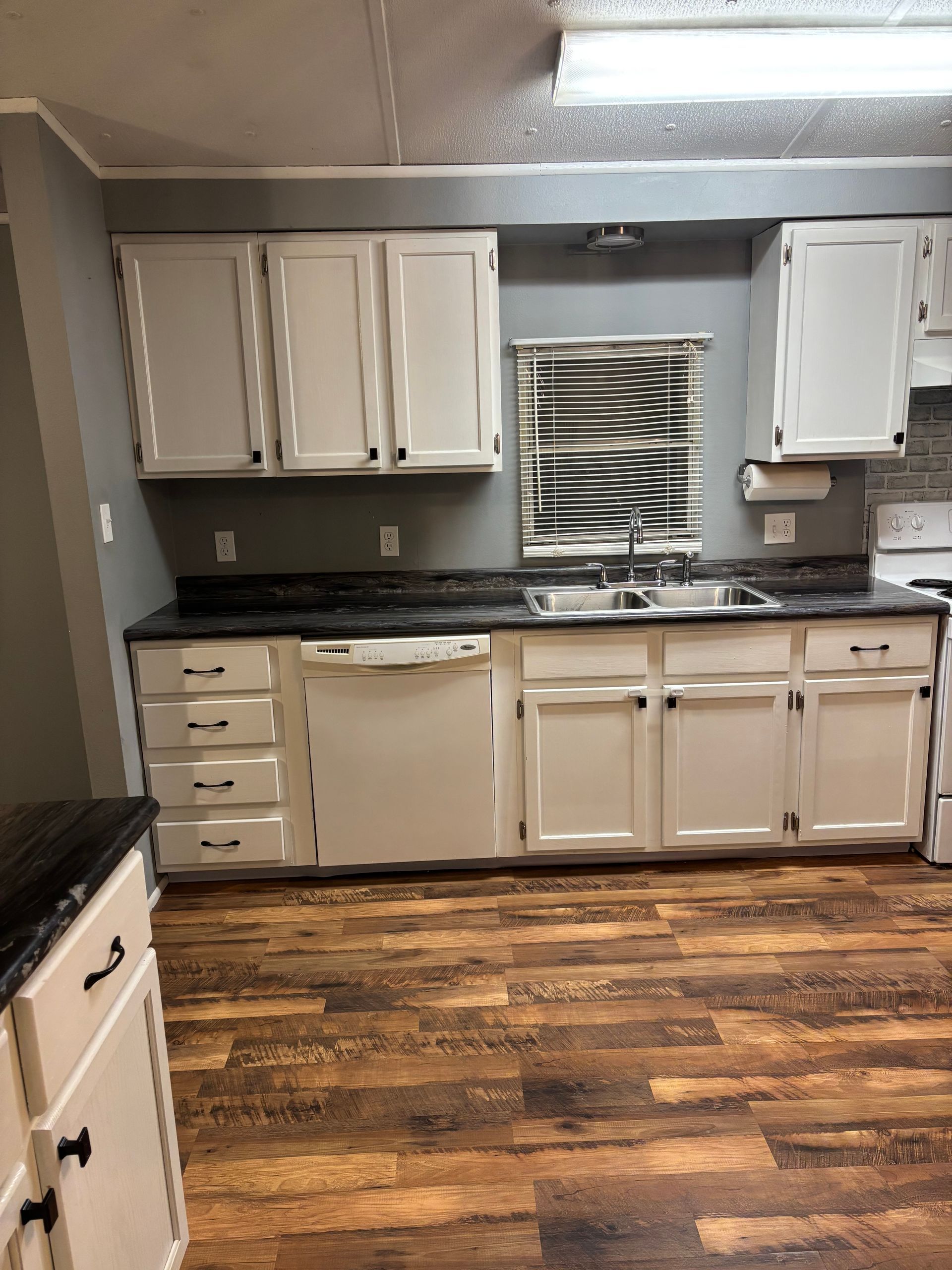 Kitchen with white cabinets, dark countertop, and wood-look flooring. Window with blinds, and silver sink.