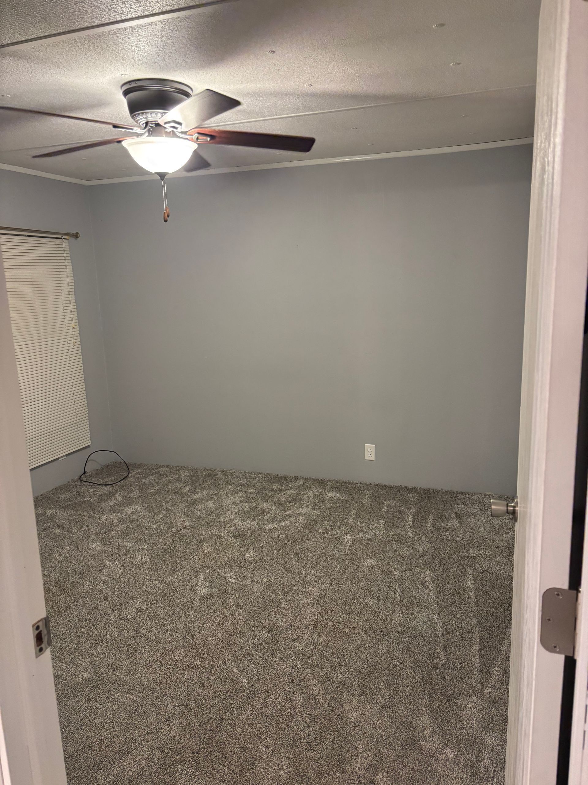 Empty bedroom with gray walls, speckled carpet, ceiling fan, and partially visible doorframe.