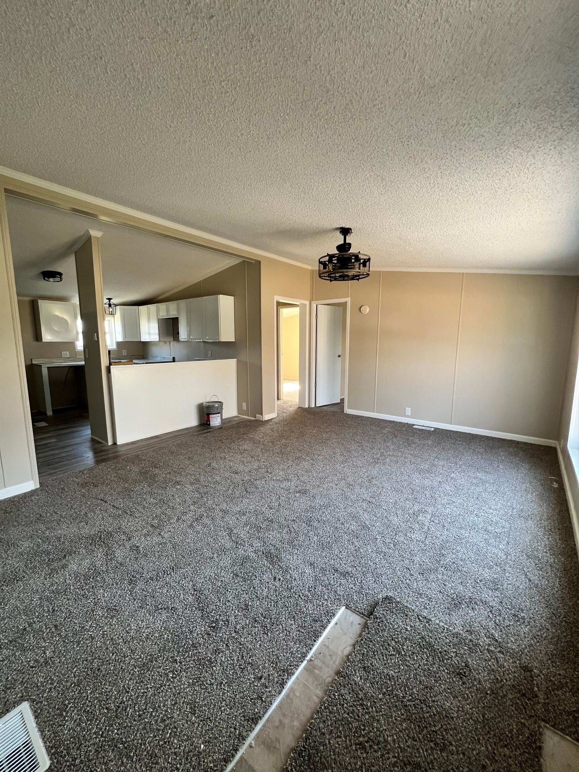 Empty living room with speckled carpet, neutral walls, and view of the kitchen and hallway.