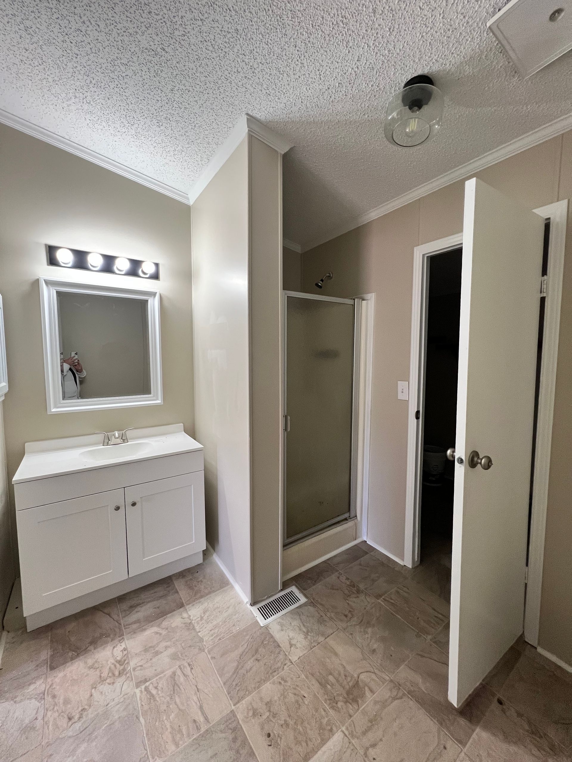 Bathroom interior with a white vanity, a mirror, shower, and open door. Beige tile floor and textured ceiling.