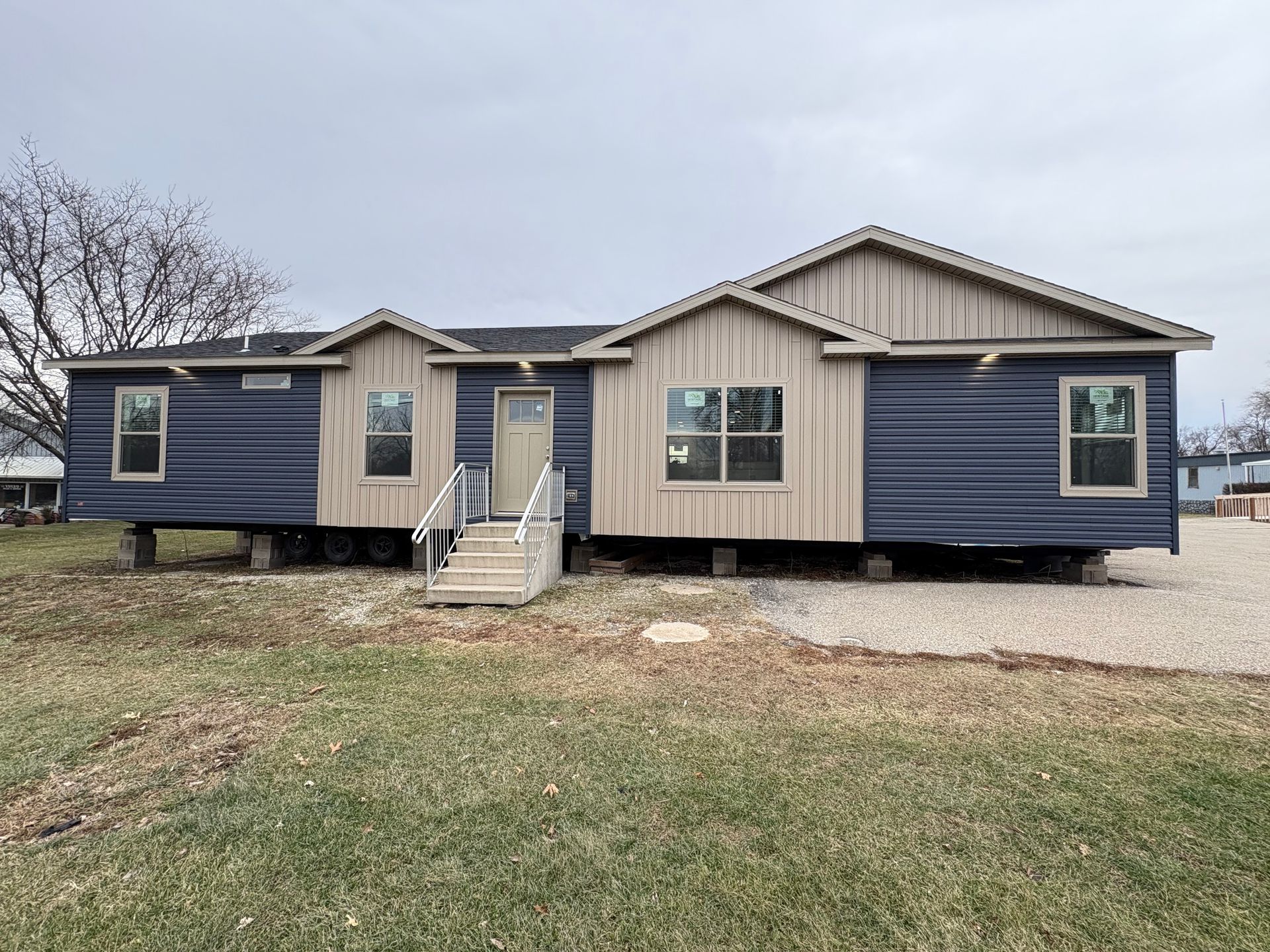 A partially constructed blue modular home with a front door and stairs, on a gravel lot under a cloudy sky.