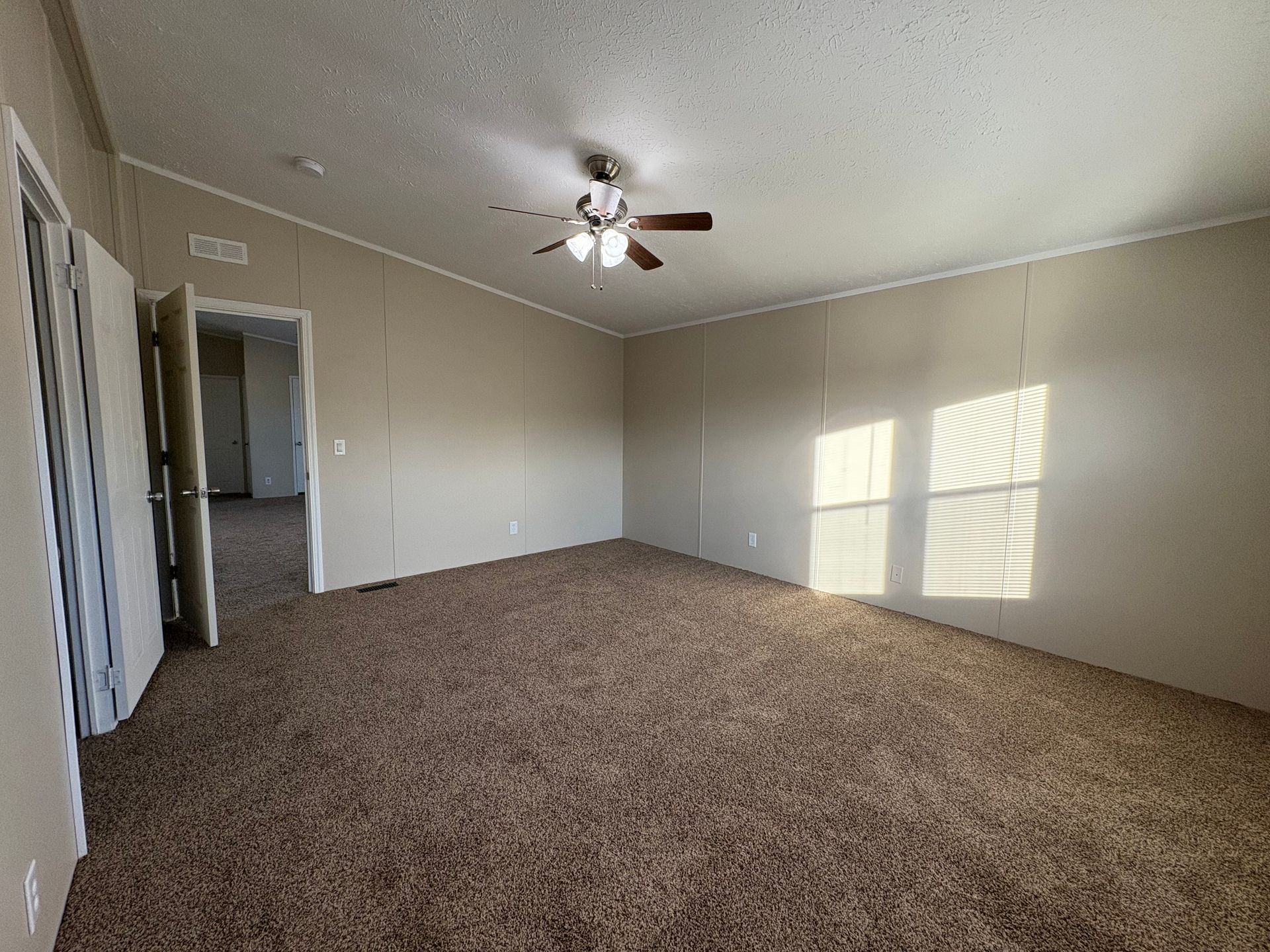 Empty room with brown carpet, tan walls, and a ceiling fan. Sunlight streams through a window.