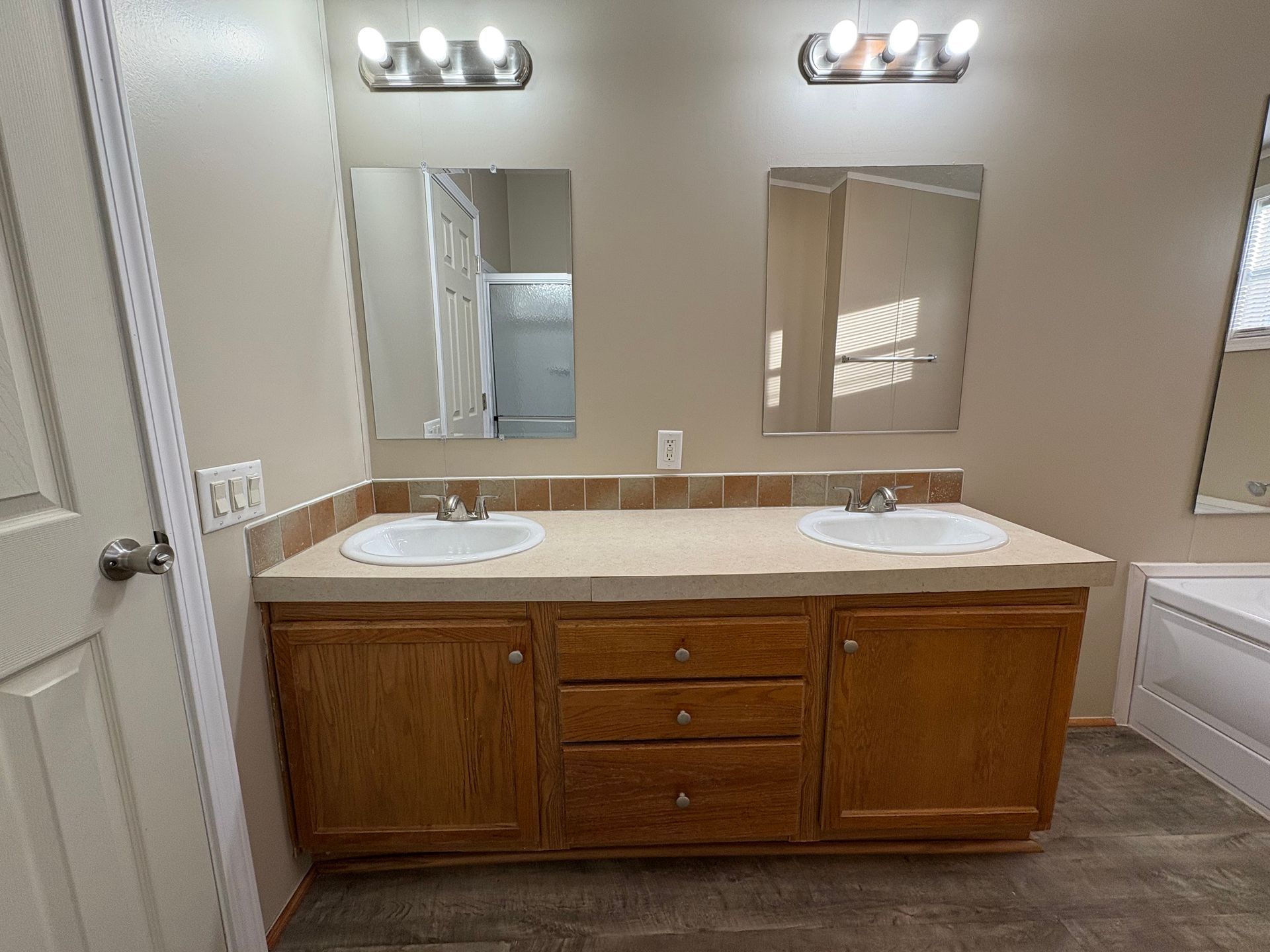 Bathroom with double vanity, mirrors, and light fixtures. Beige countertop, wooden cabinets, and gray flooring.