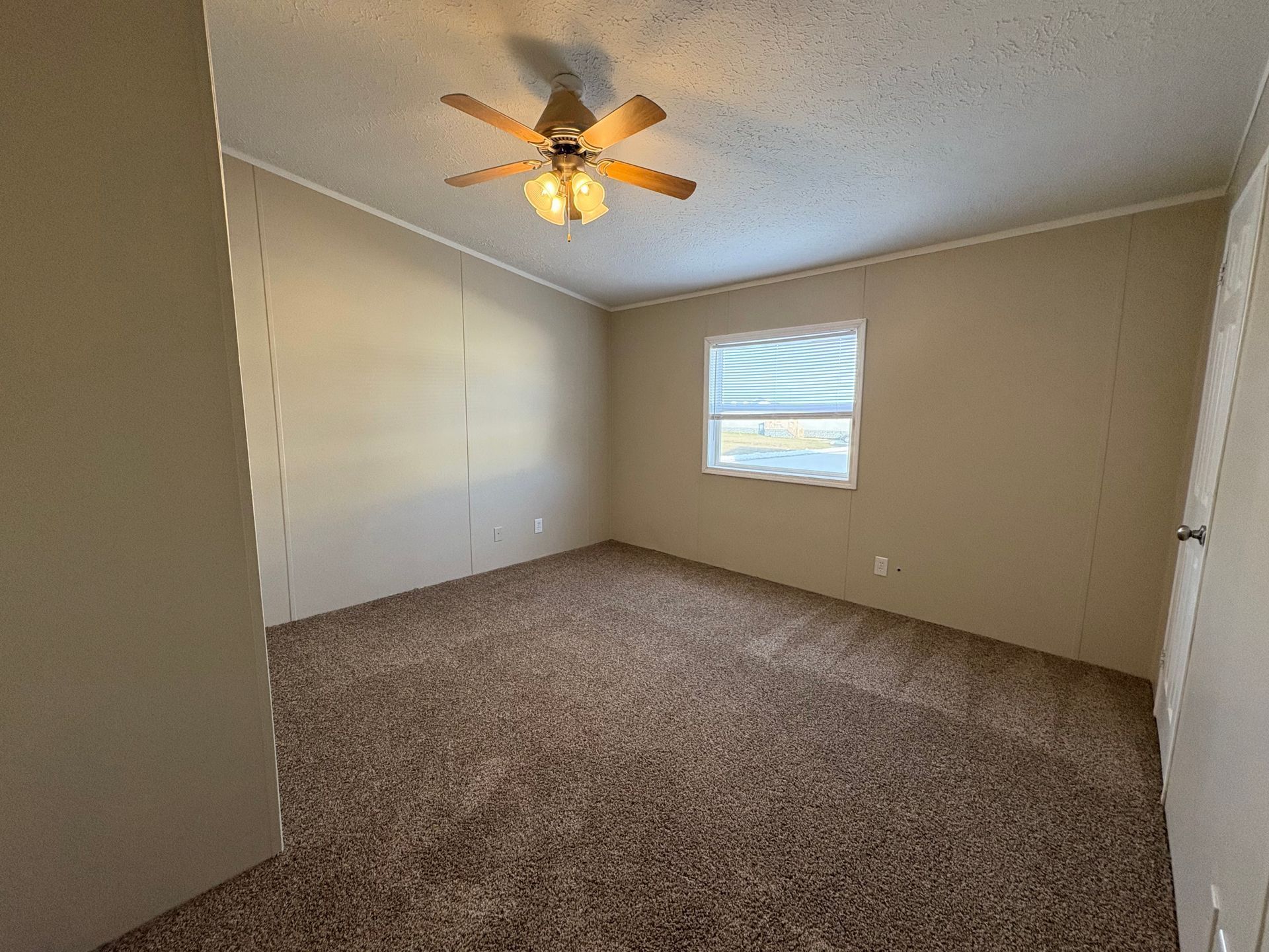 Empty bedroom with brown carpet, tan walls, ceiling fan, and window with blinds.