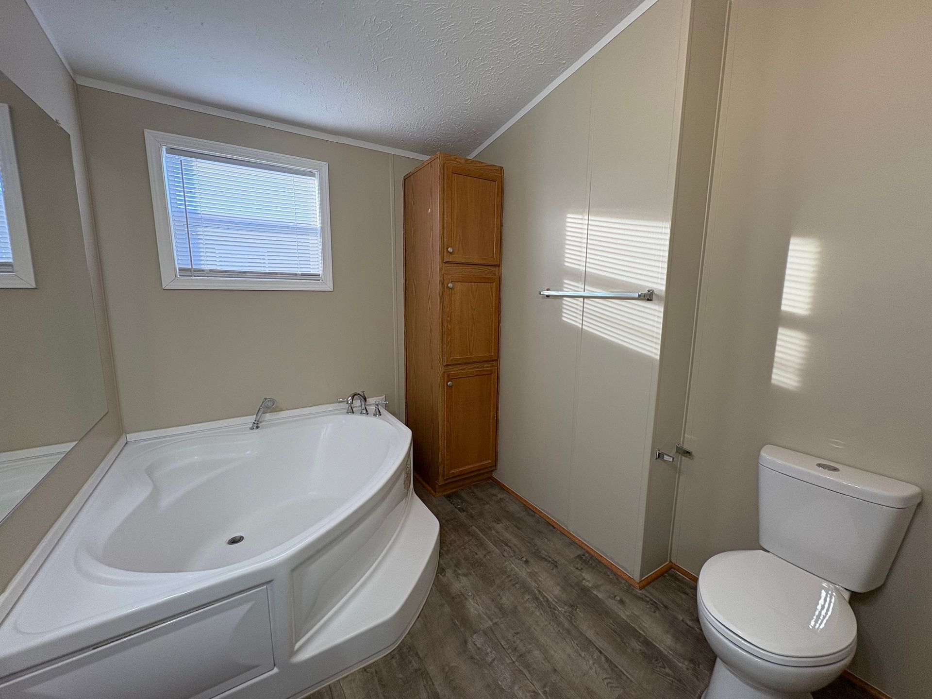 Bathroom with a white bathtub, toilet, wooden cabinet, and a window. Light beige walls and gray flooring.