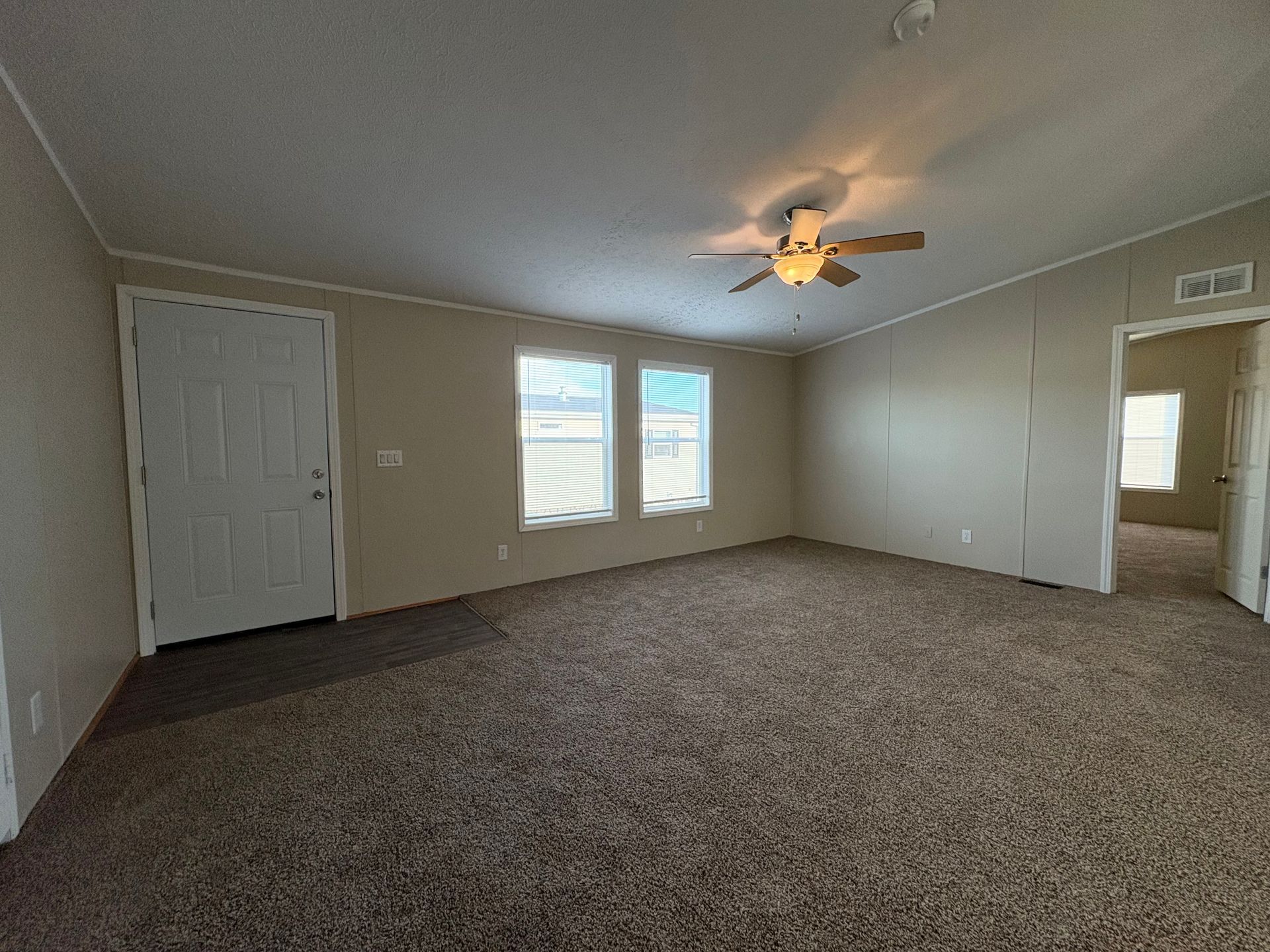 Empty living room with tan walls, brown carpet, ceiling fan, and windows.