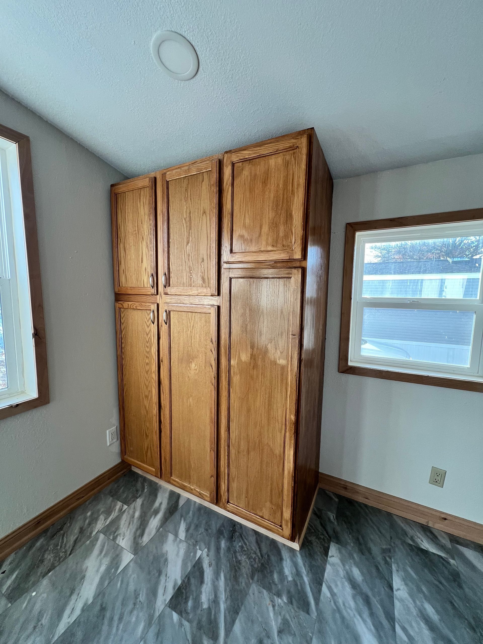 Tall wooden cabinet in a corner, between two windows in a room with wood-look flooring.