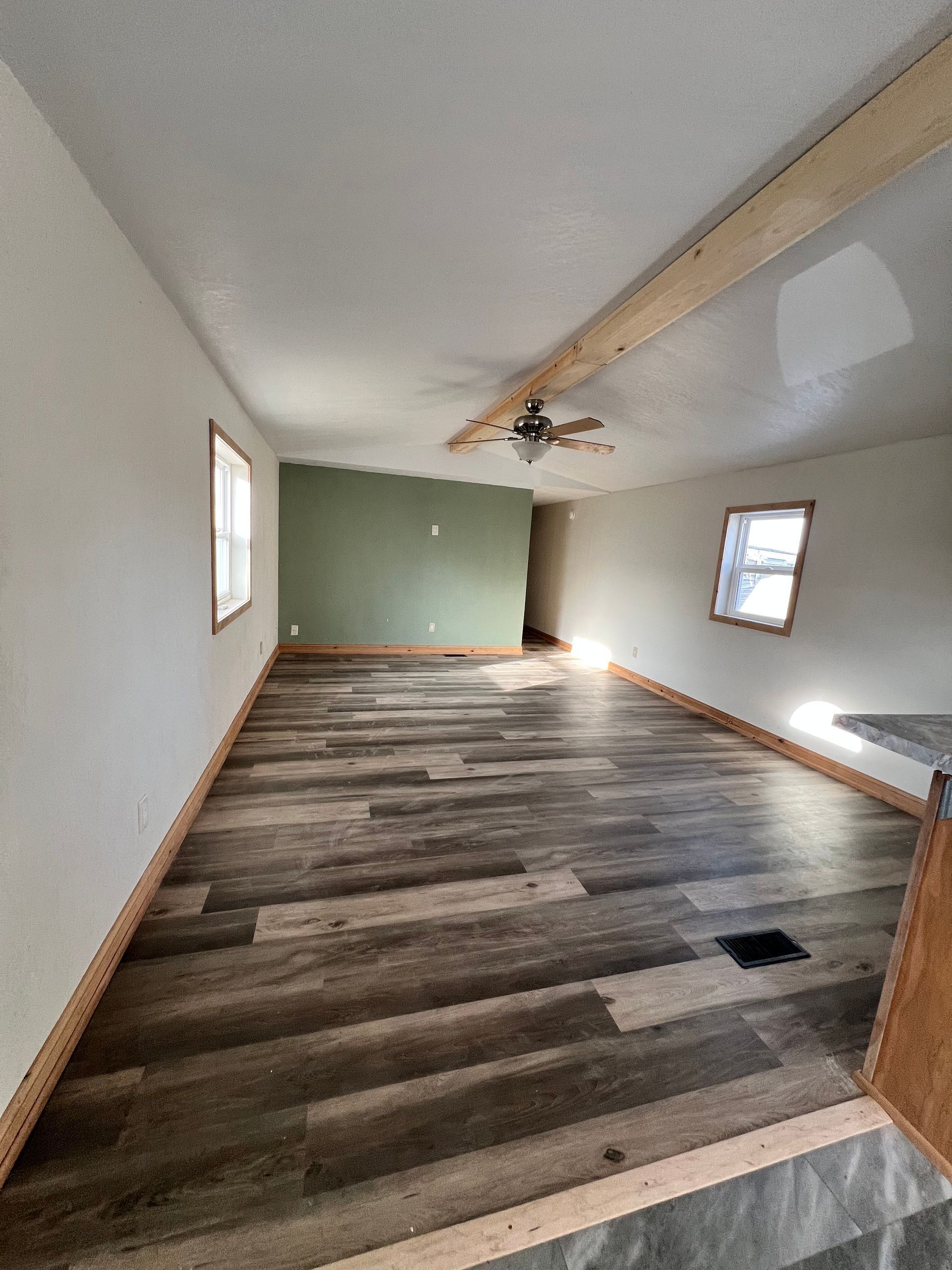 Interior view of a room with wood flooring, green accent wall, windows, and exposed ceiling beam.