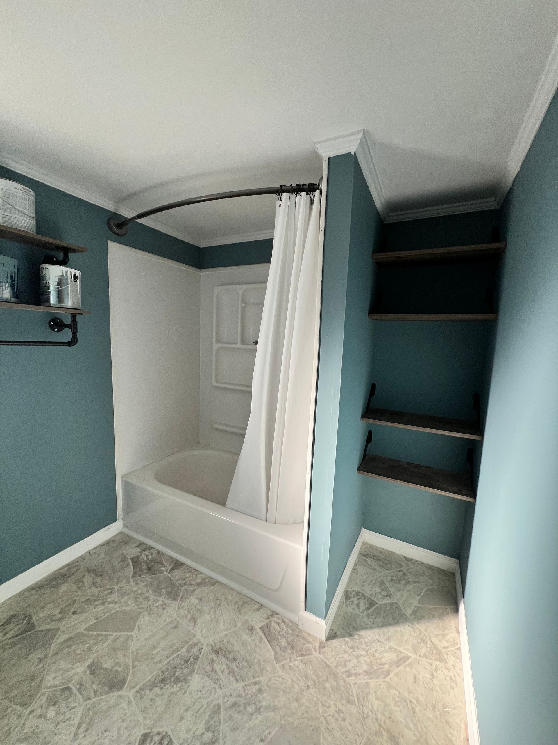 Bathroom with white bathtub, teal walls, shelves, and gray flooring.