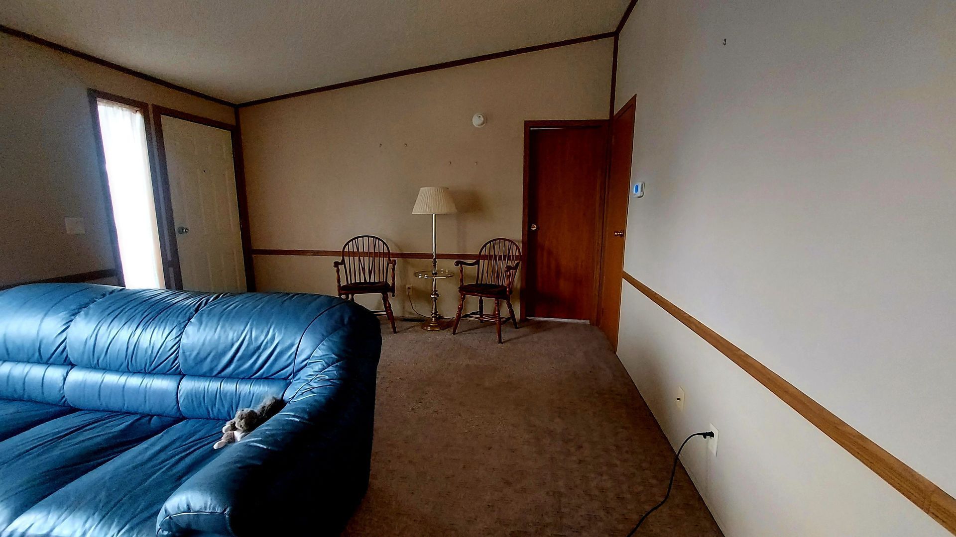 Living room with blue sofa, two chairs, a lamp, and a closed brown door. Brown carpet and beige walls.