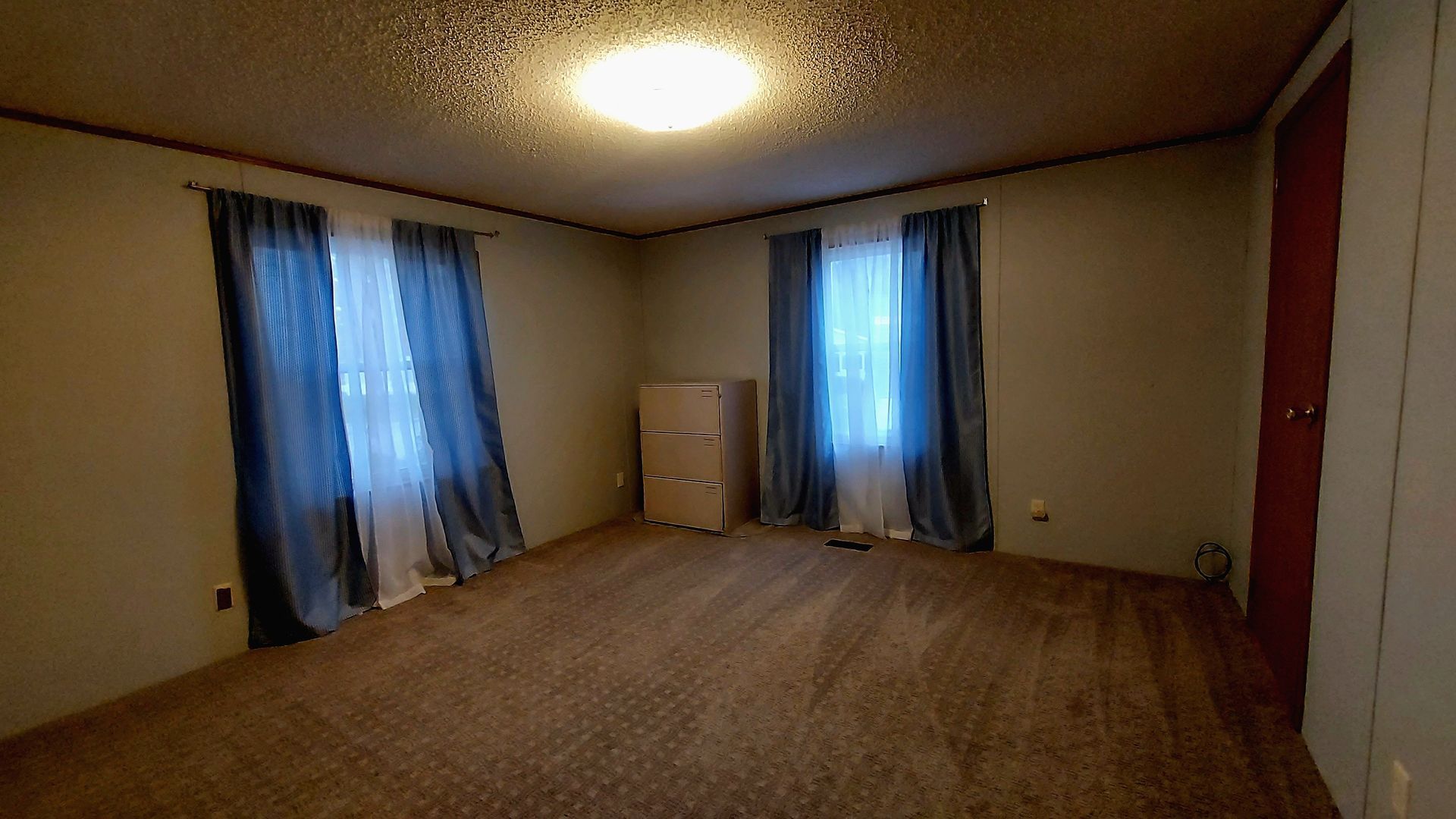 Empty bedroom with brown carpet, two windows with blue curtains, and a white cabinet.