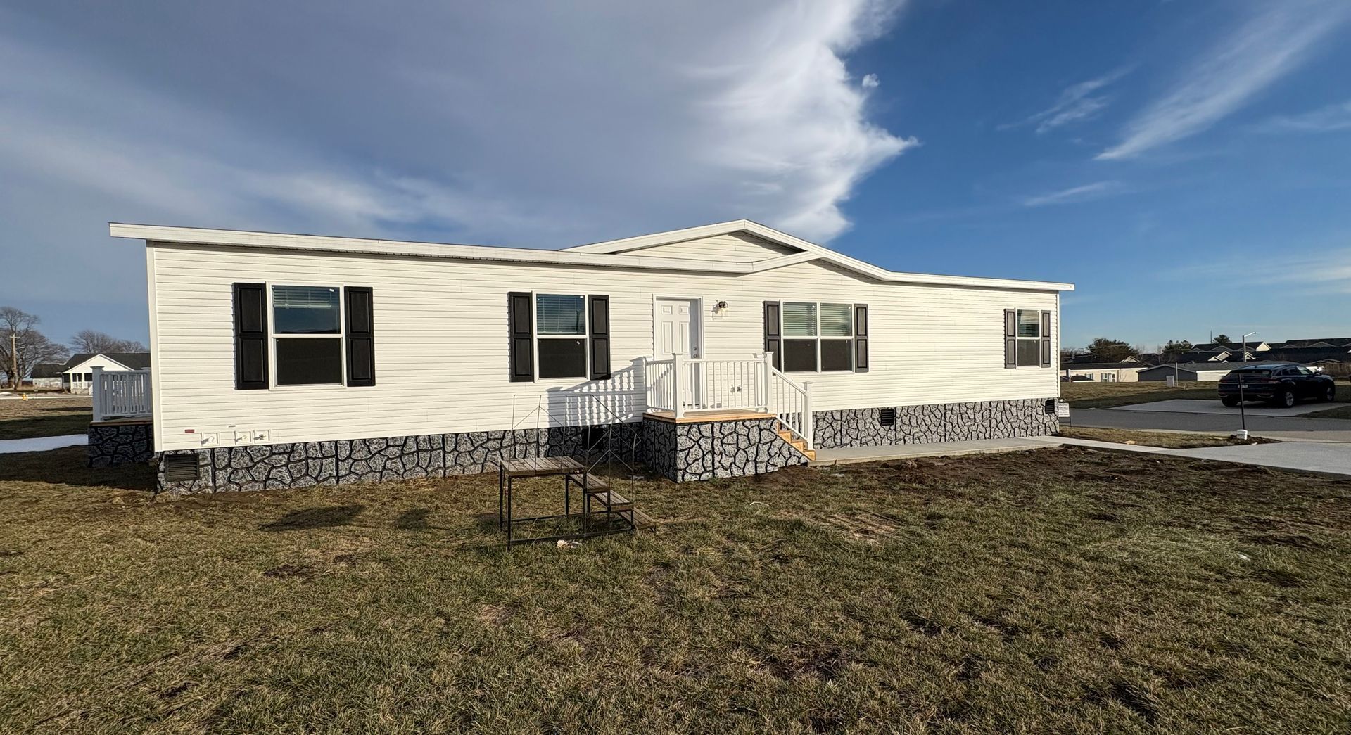 White manufactured home with dark shutters and gray foundation, set on grassy land under a blue sky.