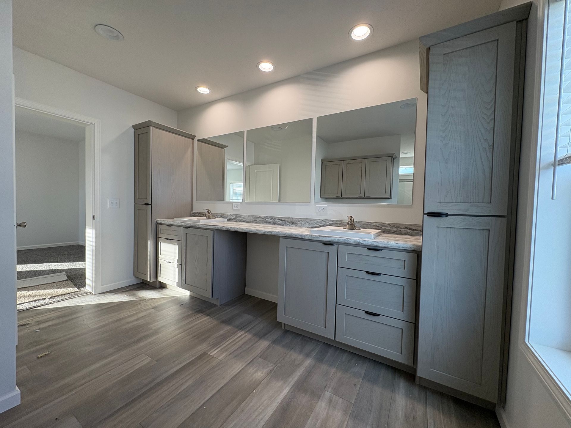 Bathroom with gray cabinetry, mirrors, and wood-look flooring. Light fixtures in ceiling, window in the right.