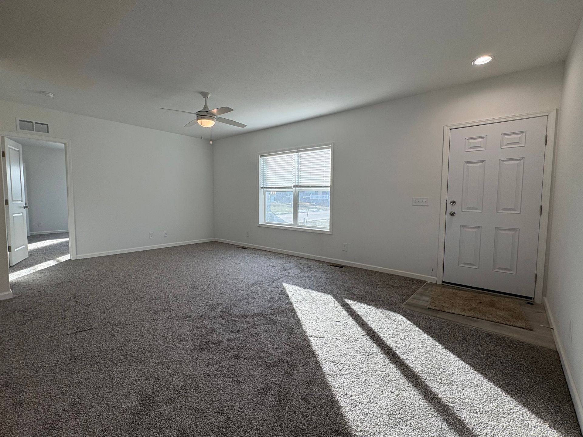 Empty living room with gray carpet, white walls, a closed door, and a window.