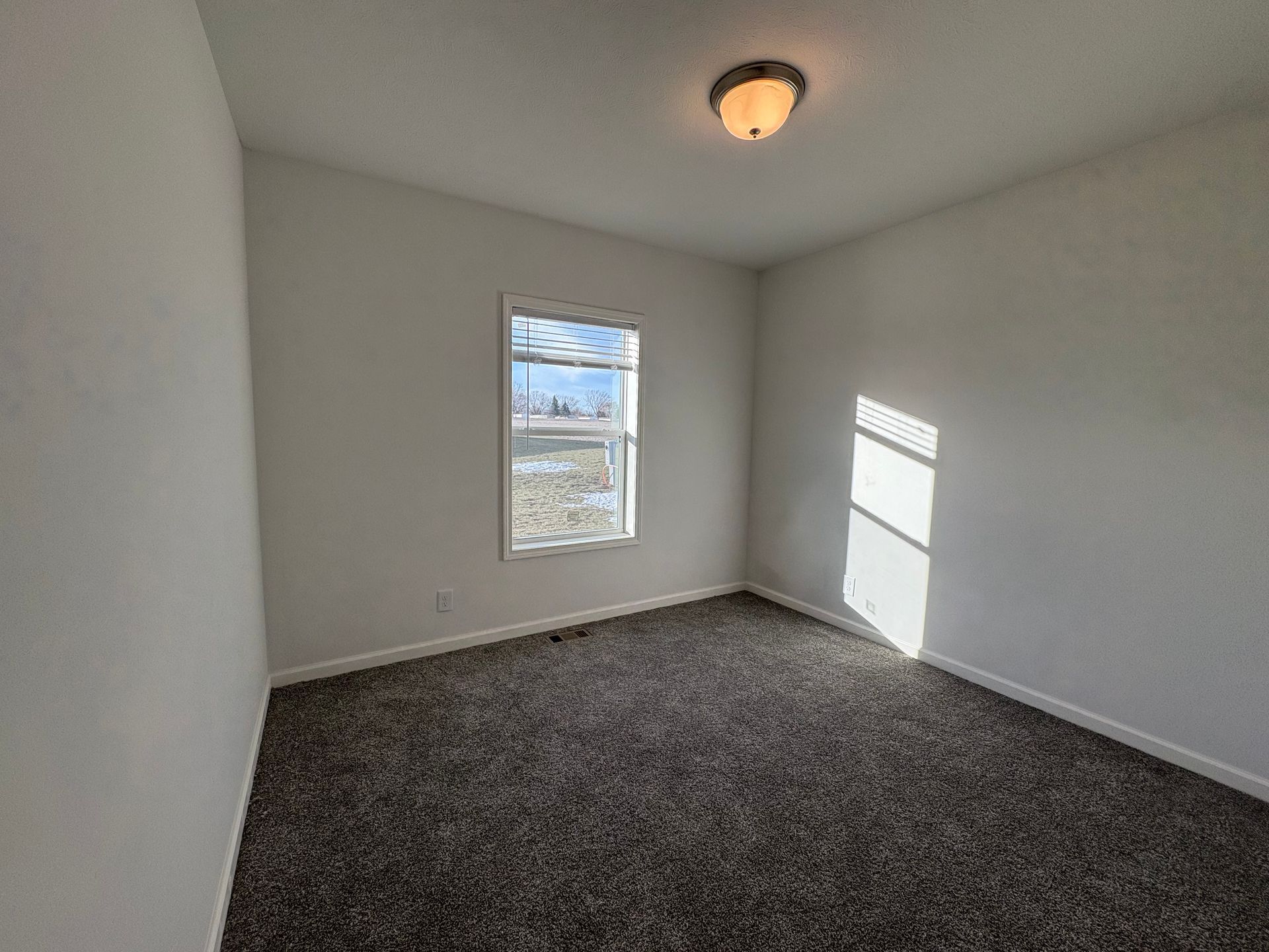 Empty bedroom with white walls, window, and dark carpet. Ceiling light visible.