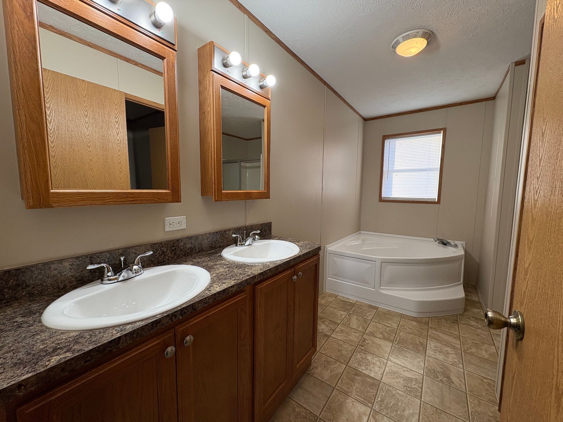 Bathroom with dual sinks, mirrors, tub, window, and brown cabinets and walls.