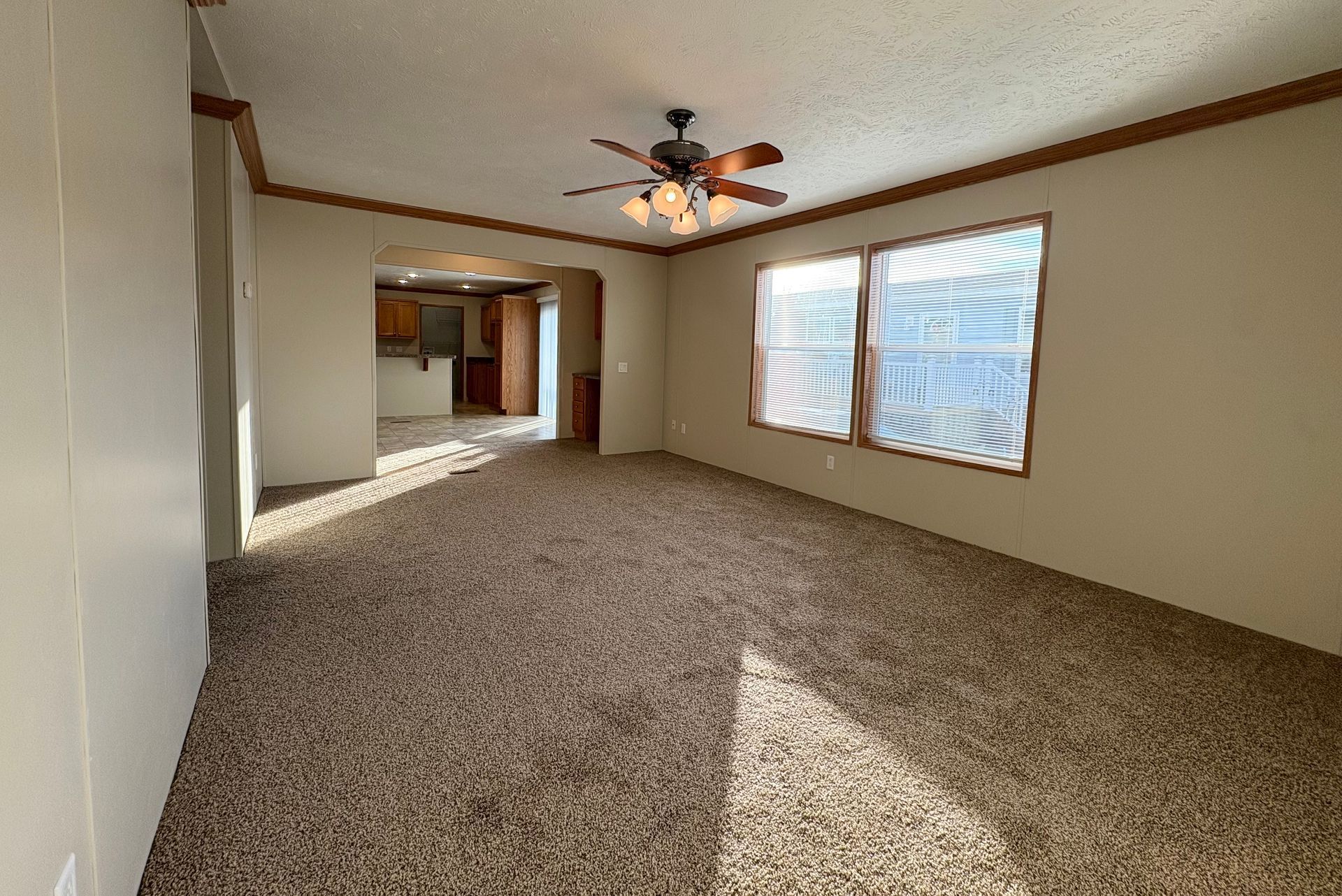 Empty living room with tan carpet, beige walls, and a view into the kitchen.