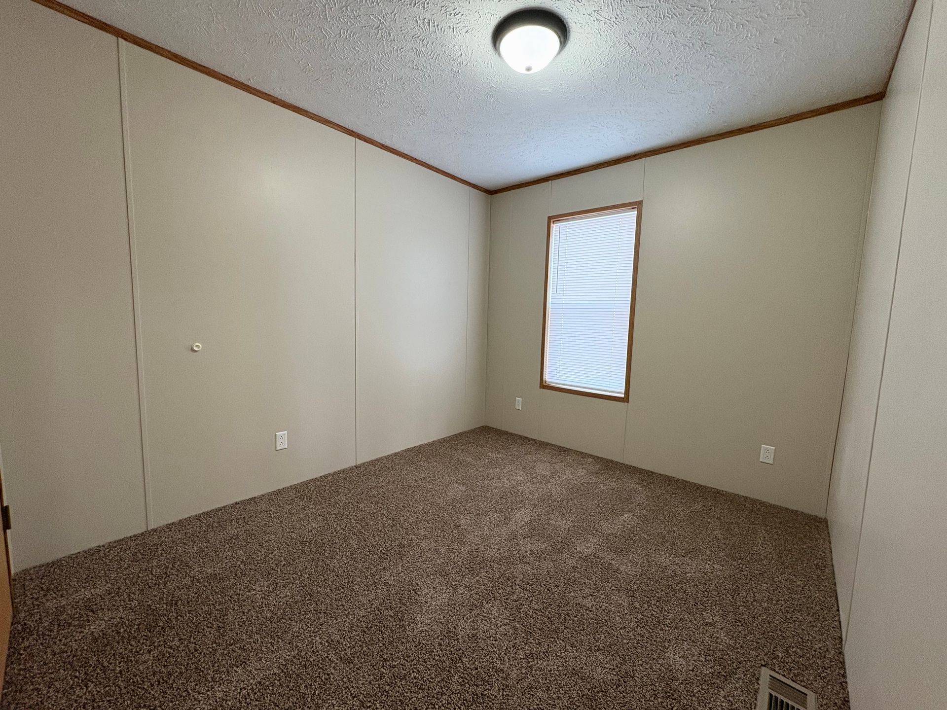 Empty room with brown carpet, tan walls, small window with blinds, and ceiling light.