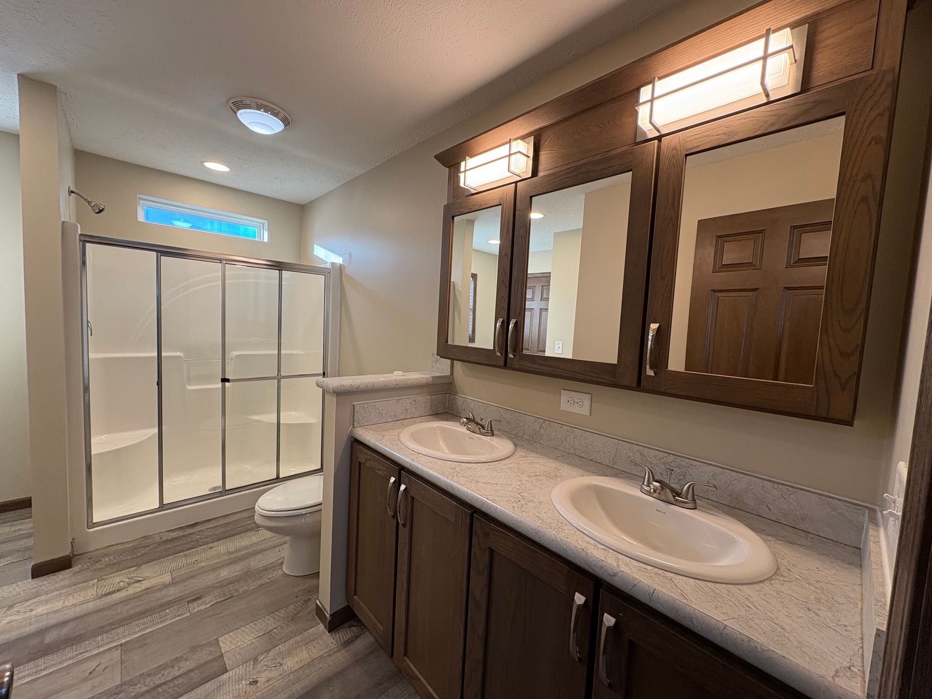Bathroom with double vanity, mirrors, shower, and toilet. Features brown cabinets, gray floor, and white countertops.