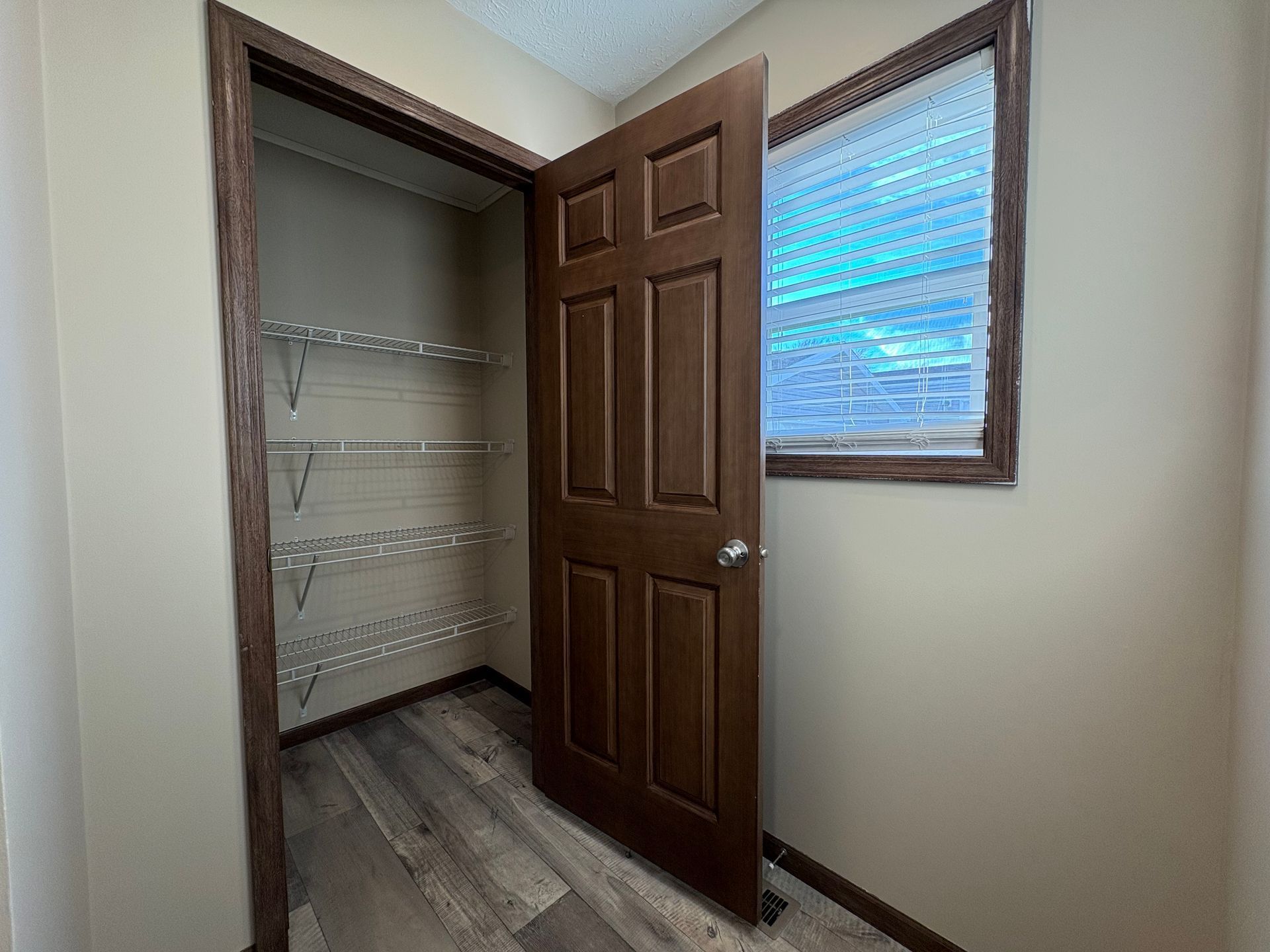 Closet with shelves, open brown door, window on right. Gray shelves and flooring. Beige walls.