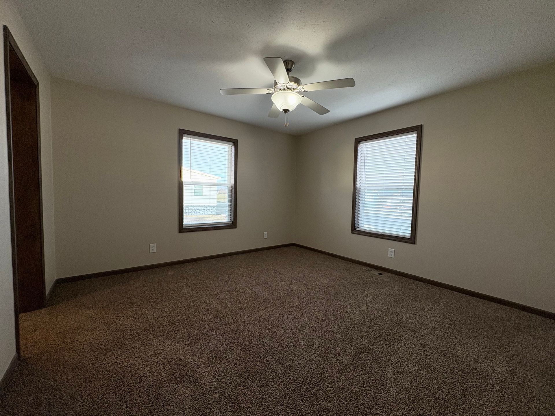 Empty bedroom with brown carpet, two windows, and a ceiling fan.