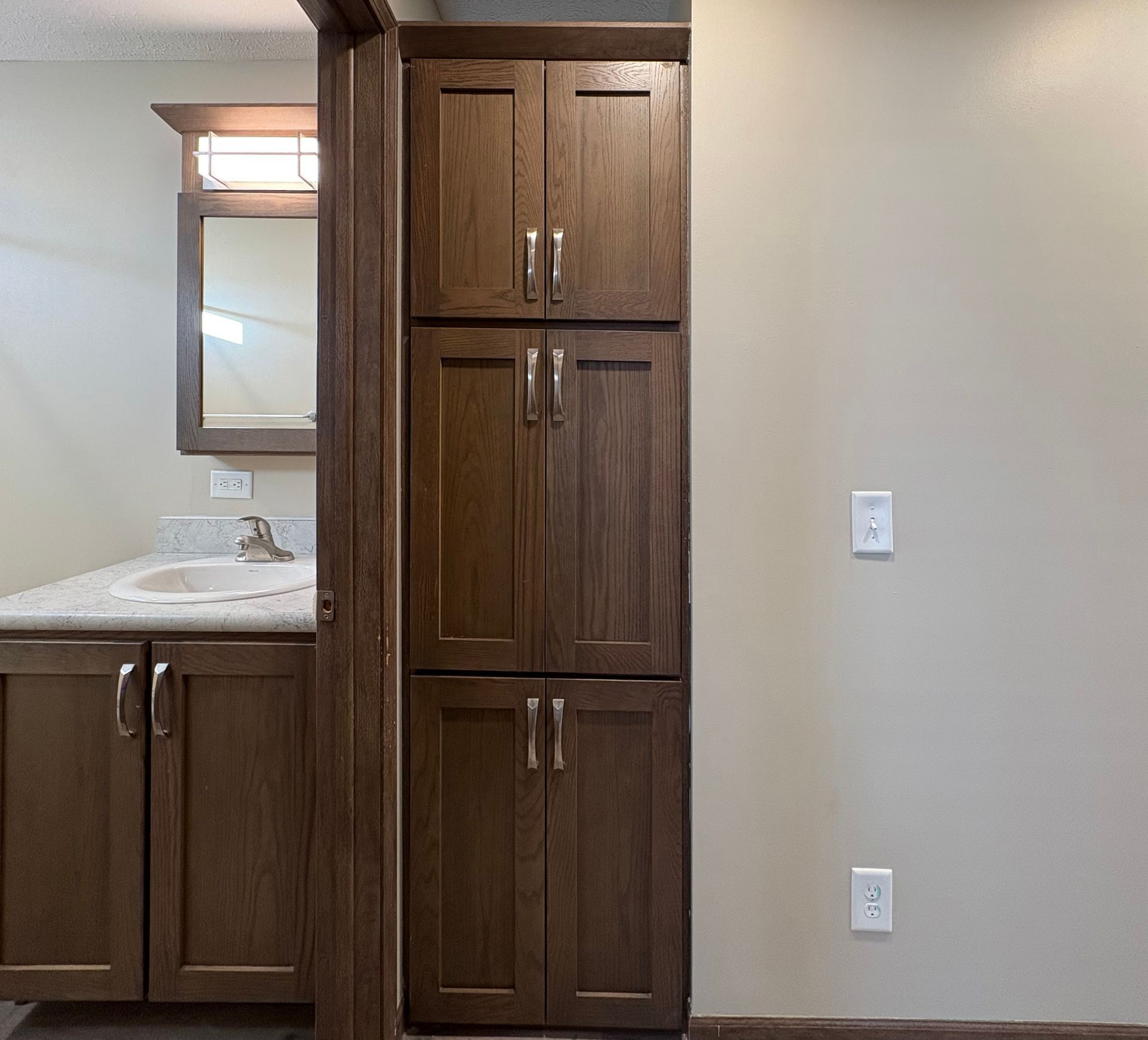 Bathroom with wood cabinets, light countertop, and a tall storage cabinet. Beige walls.
