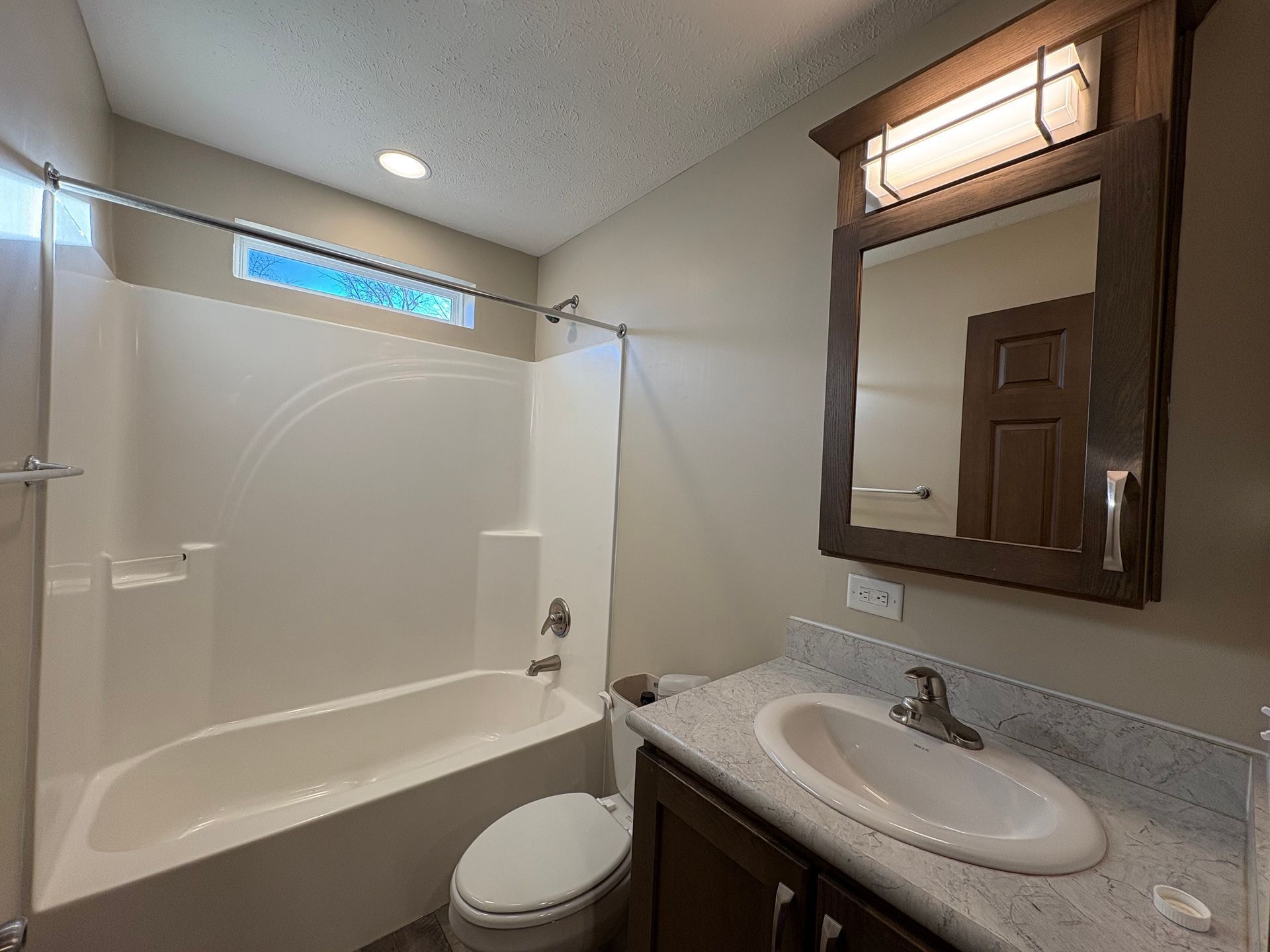 Bathroom with tub, toilet, vanity with mirror. Neutral tones, overhead lighting, window above tub.