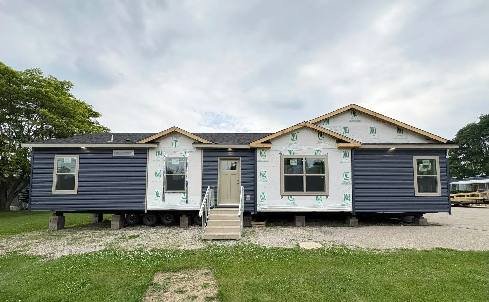 A partially constructed blue modular home with a front door and stairs, on a gravel lot under a cloudy sky.