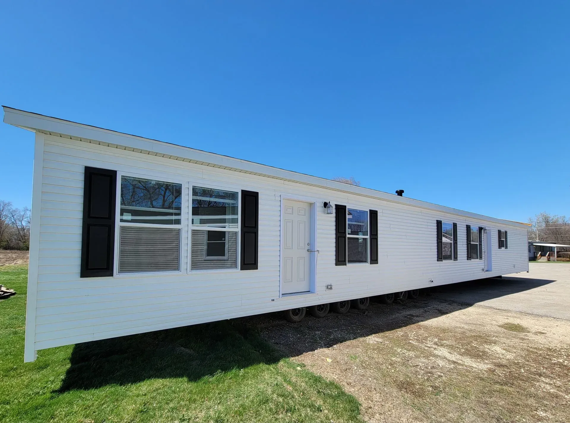 White mobile home with black shutters, on grass and gravel, against a blue sky.