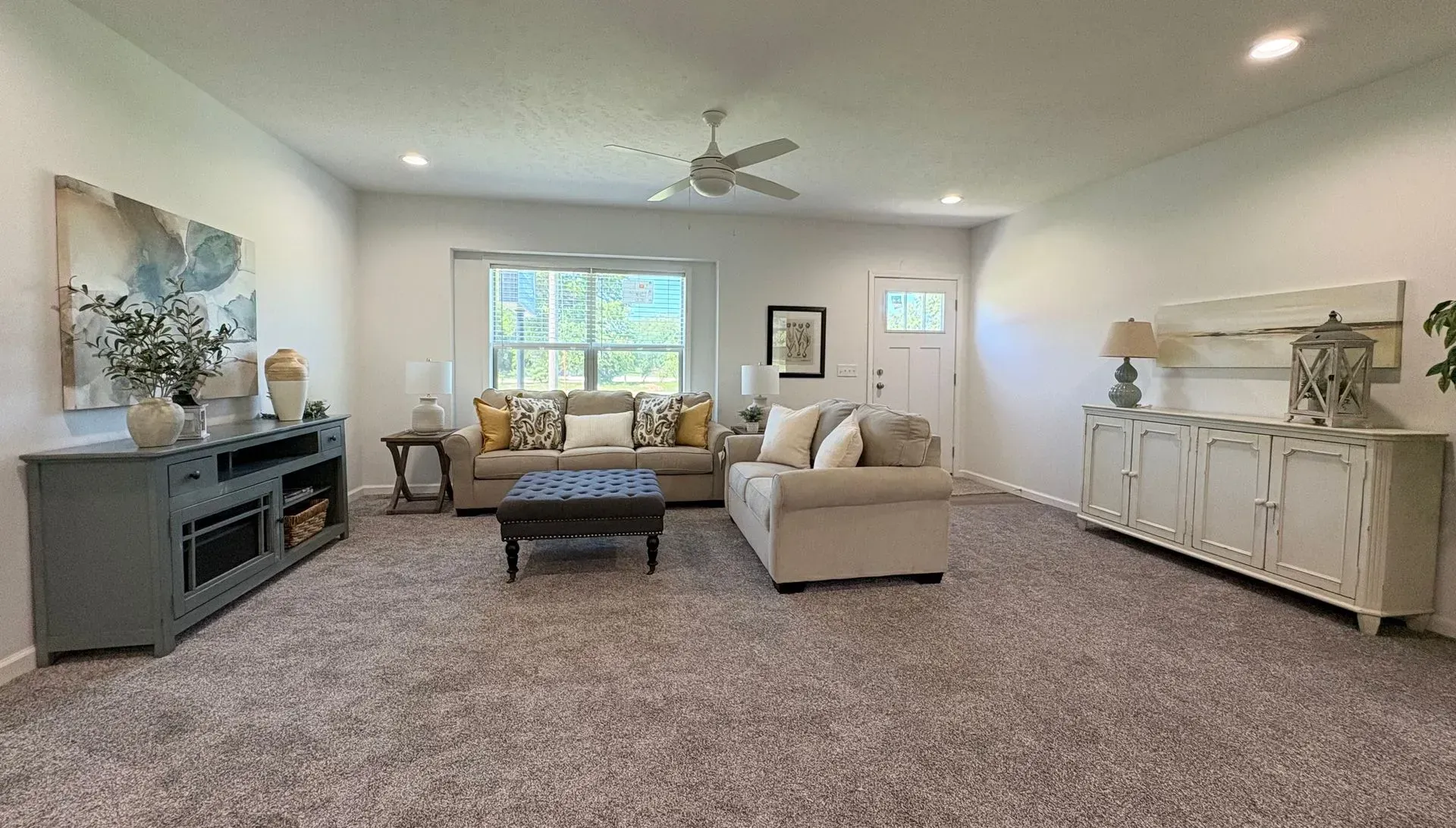Spacious living room with tan sofas, a coffee table, and large windows. Neutral color scheme and plush carpet.