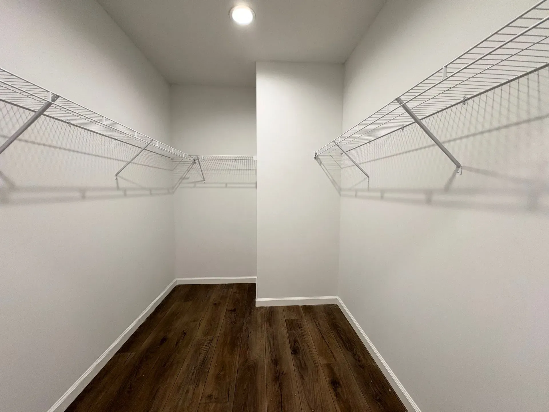 Empty walk-in closet with wire shelving on white walls, dark wood floor, and a single ceiling light.