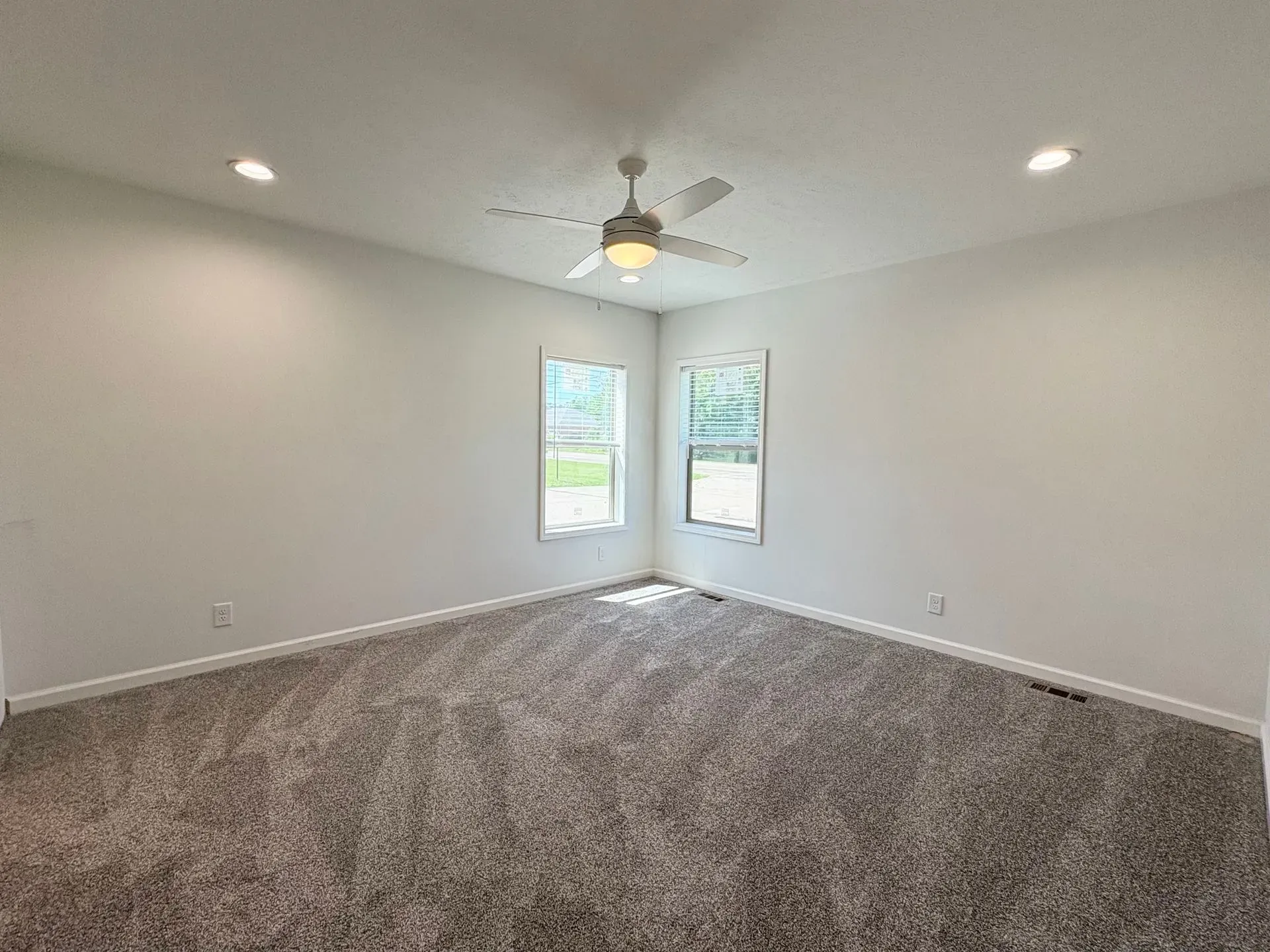 Empty bedroom with light gray carpet, white walls, two windows, and a ceiling fan.