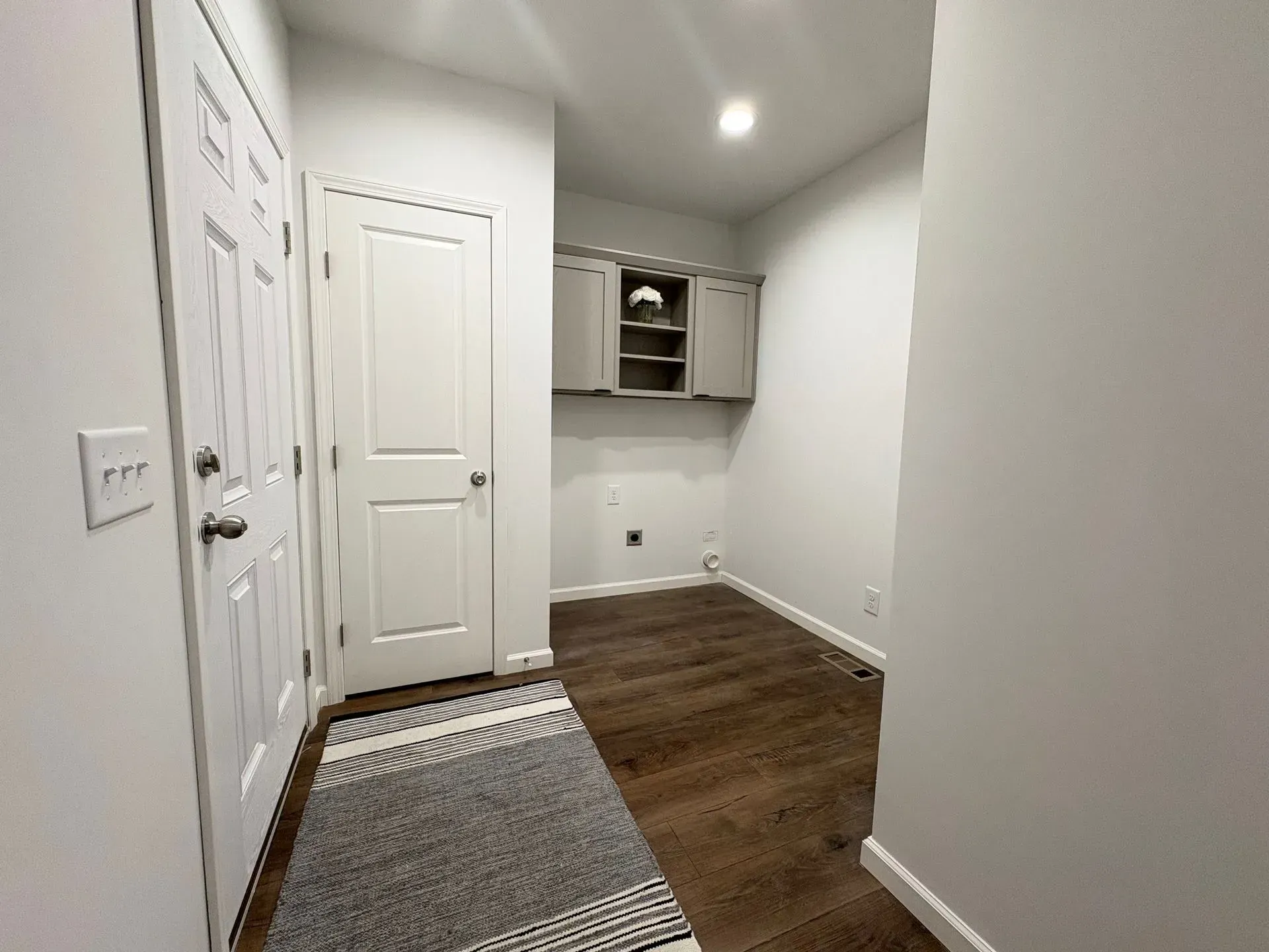 Laundry room with dark wood floor, white walls, and gray cabinets. Rug on the floor.