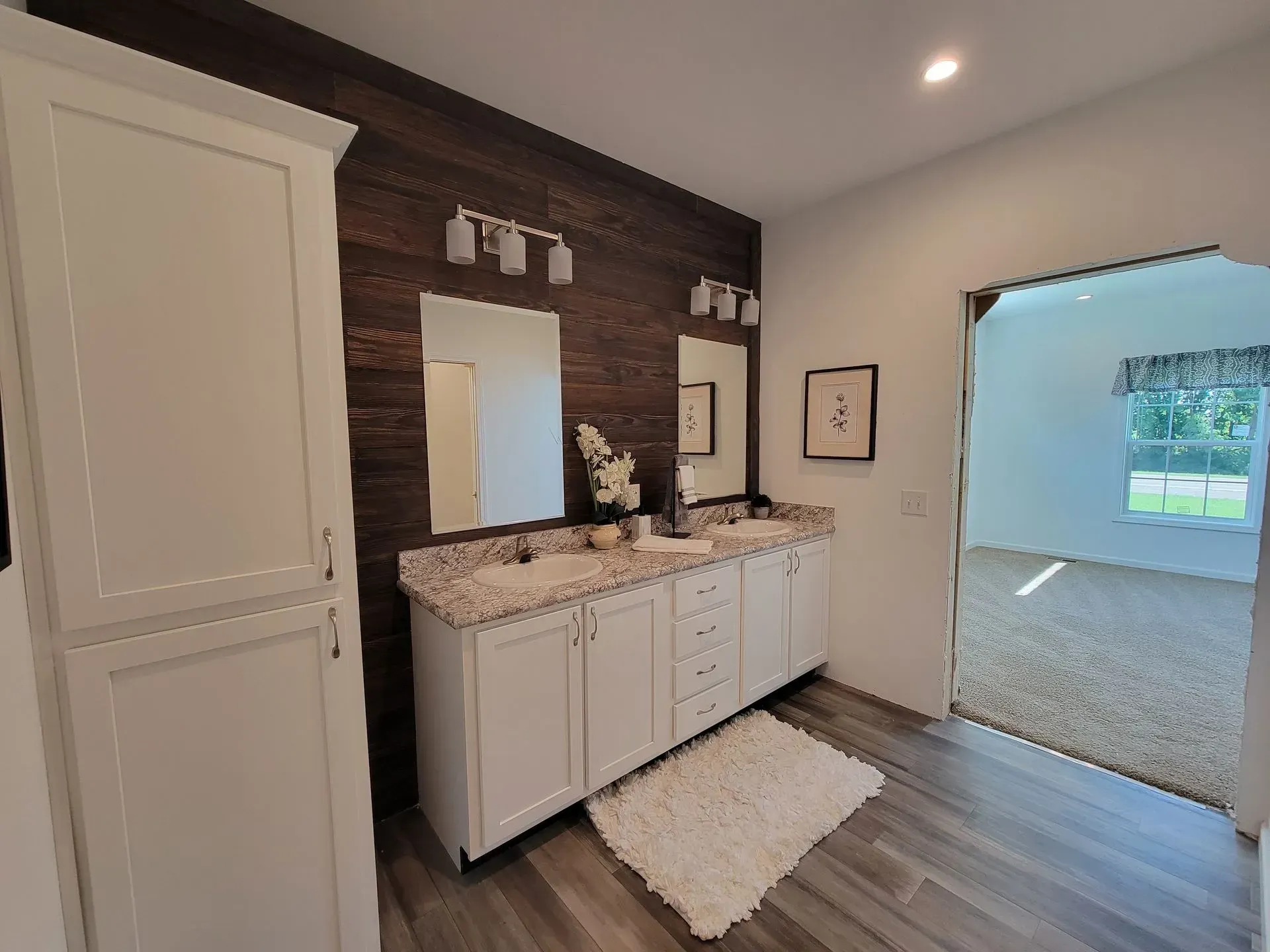 Bathroom with white cabinets, wood accent wall, dual sinks, and a doorway to a bedroom.