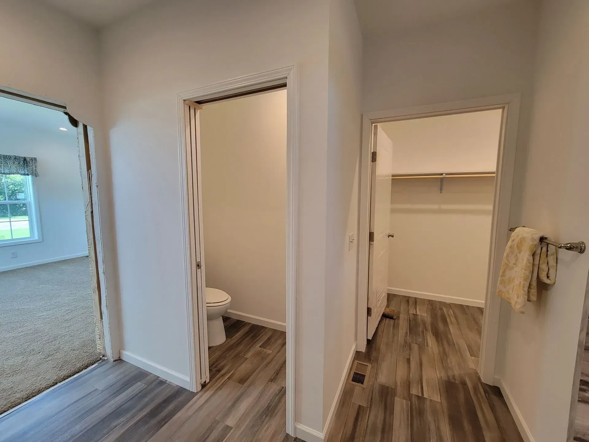 Bathroom with toilet, closet, and doorway to a bedroom, wood-look flooring, and white walls.