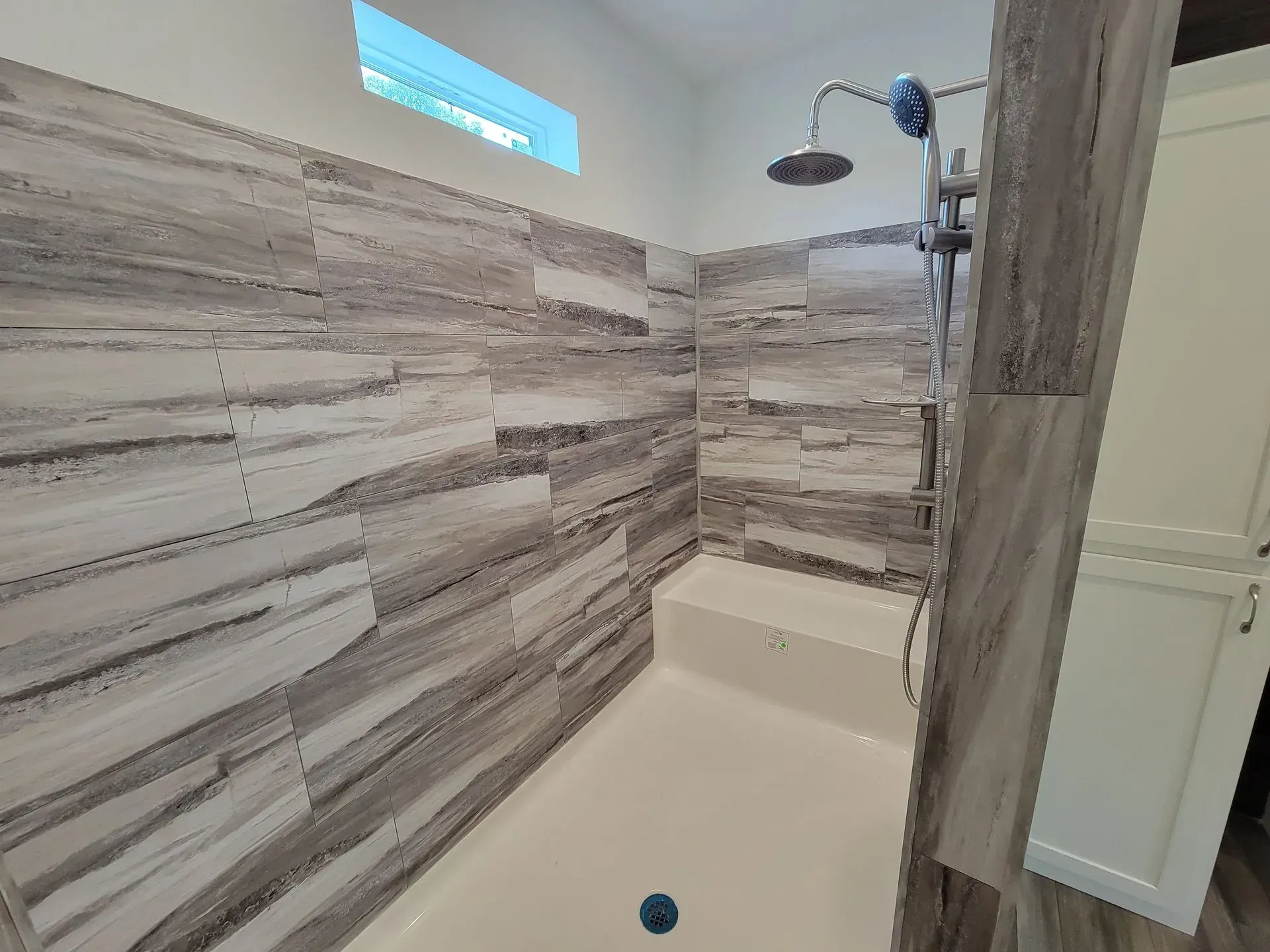 Bathroom shower with gray and white tiled walls, shower head, bench, and a small window.