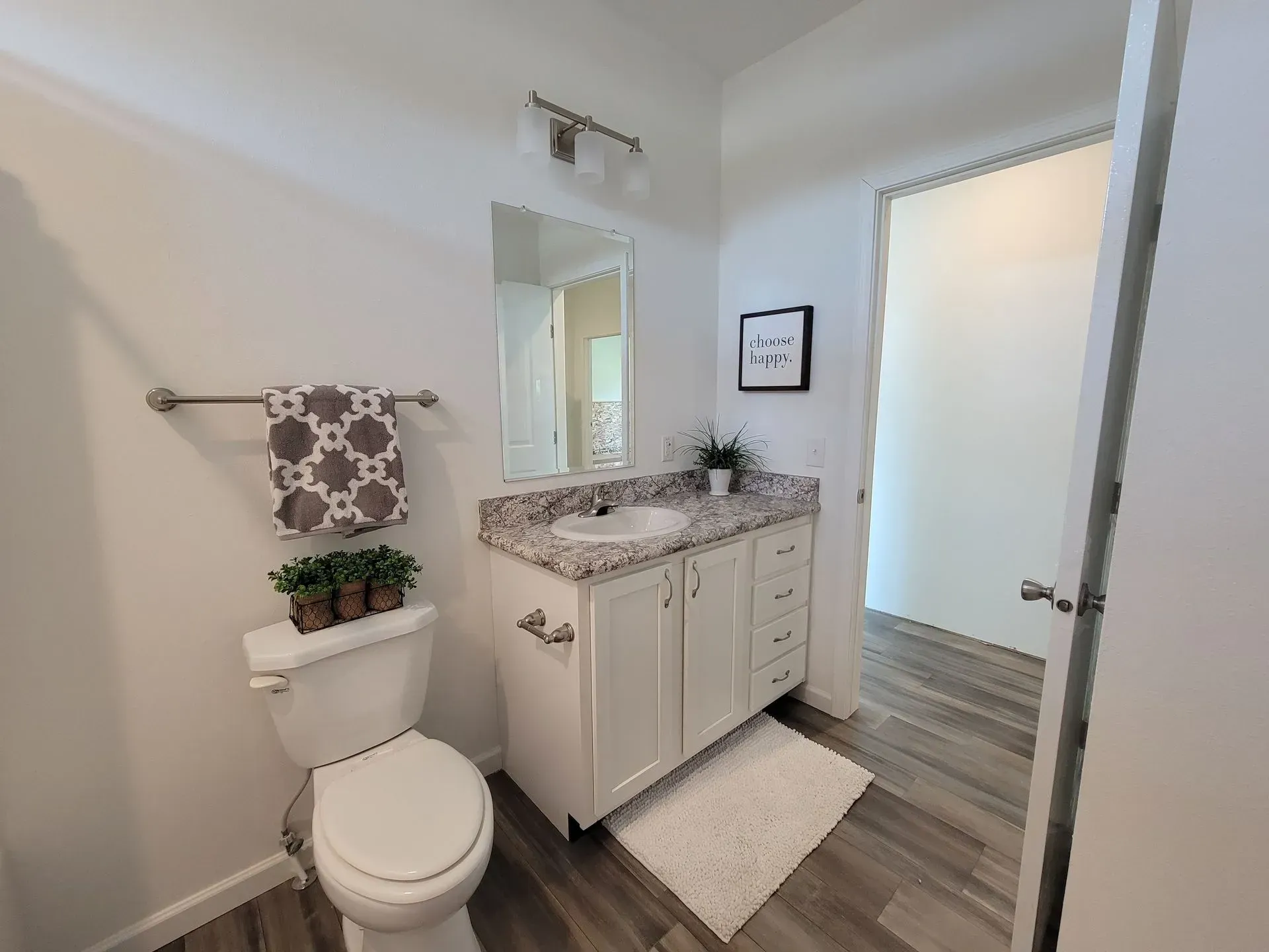 Bathroom with white vanity, toilet, and fluffy rug; wooden floor and open doorway.