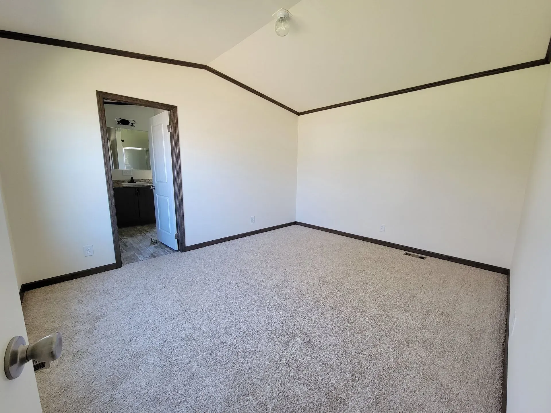 Empty room with beige carpet, white walls, dark brown trim, and an open doorway to a bathroom.
