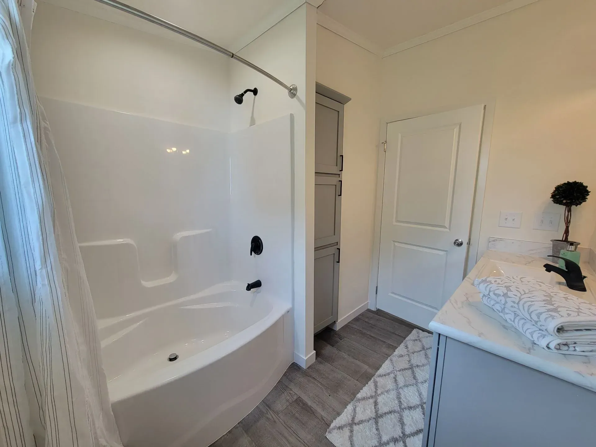 Bathroom with white tub/shower, grey cabinet, white door, and patterned rug on a wood-look floor.