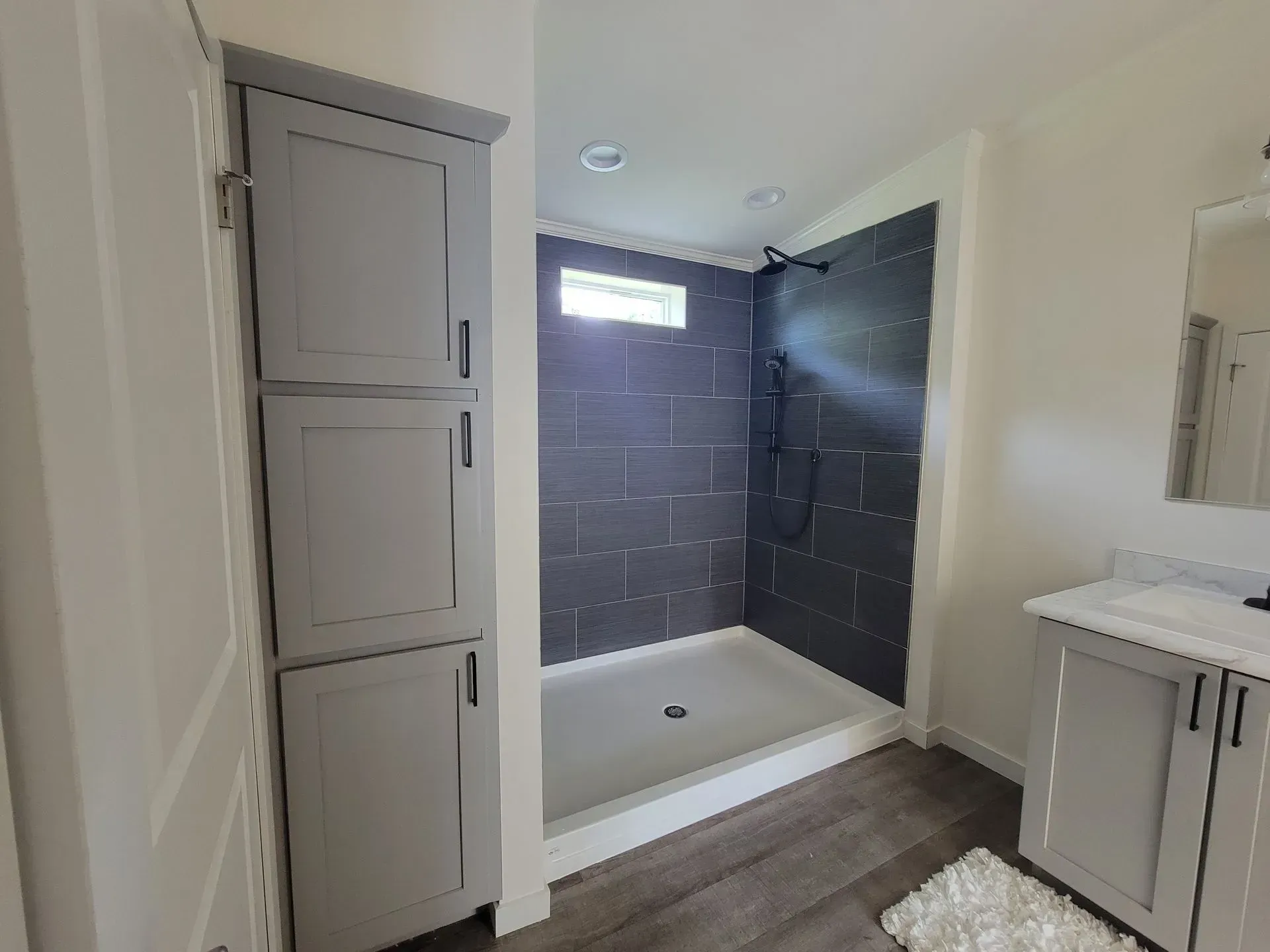 Bathroom with gray tiled shower, storage cabinet, vanity, and wood-look flooring.