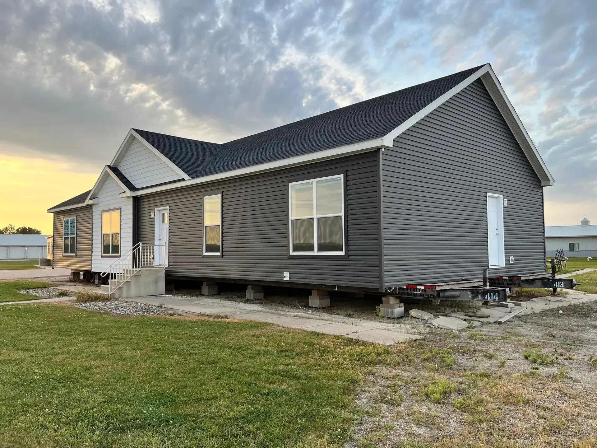 Dark gray modular home on blocks, next to a grassy area, under a cloudy sky.