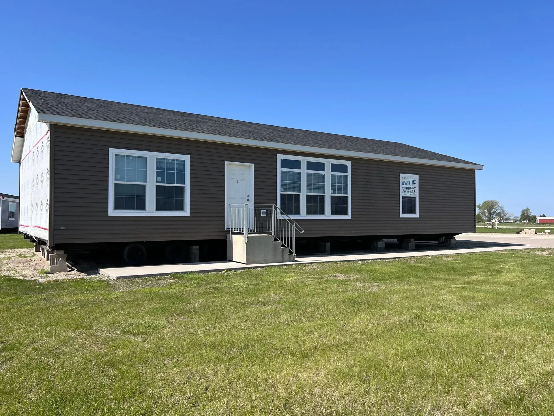 Brown-sided single-story house with white trim, door, and windows. Set on a foundation in a grassy field under a blue sky.