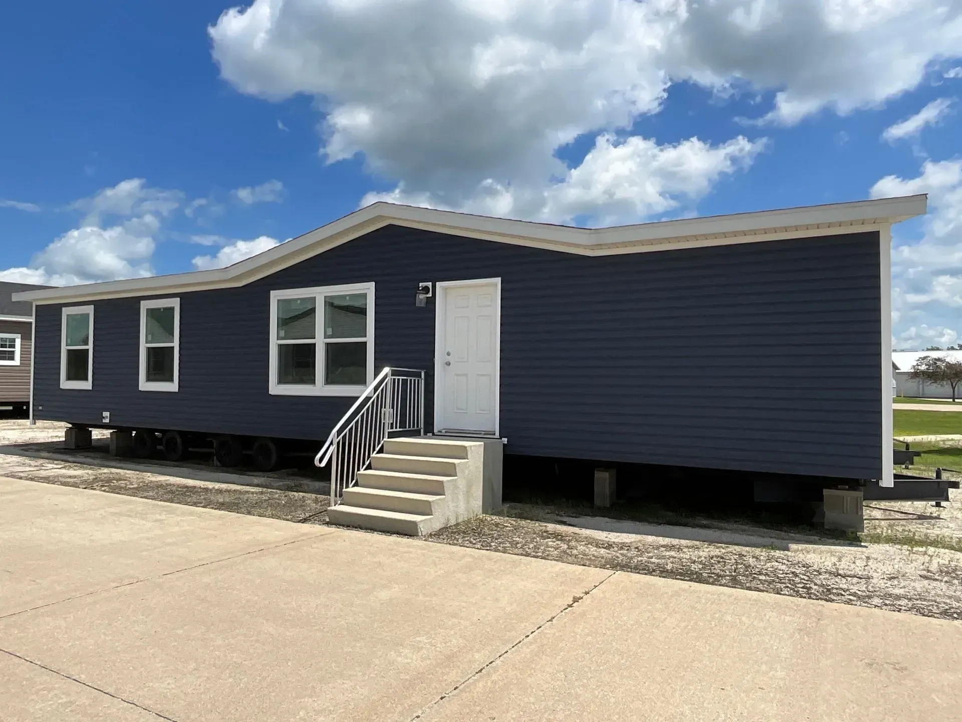 Blue-sided mobile home with white trim and a concrete entrance, parked on a paved lot under a cloudy sky.