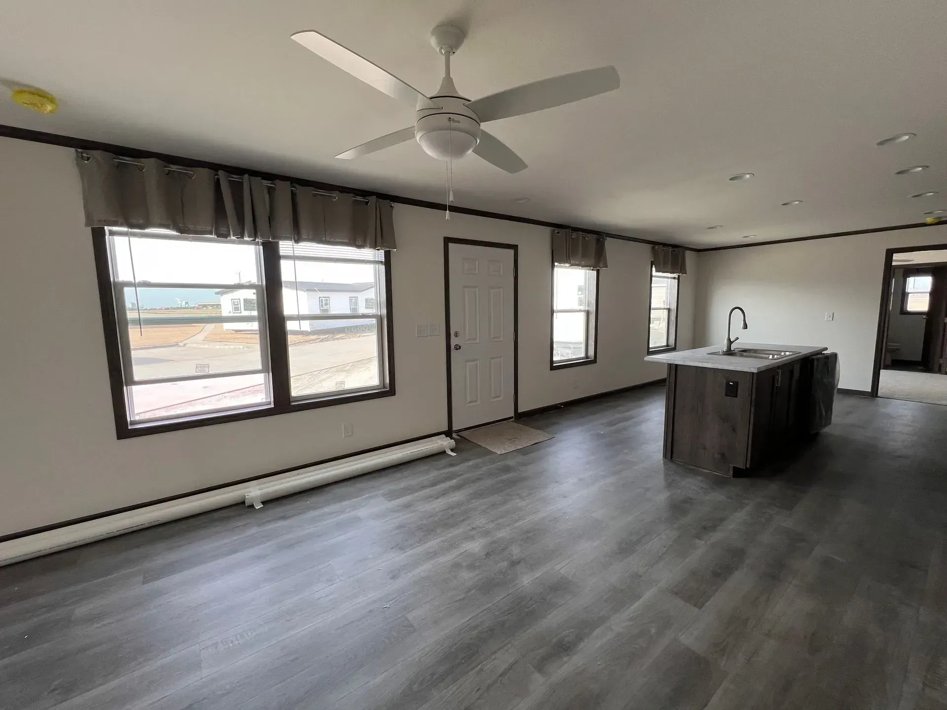 Interior view of a modern living area with grey flooring, white walls, and a ceiling fan. Includes kitchen island.