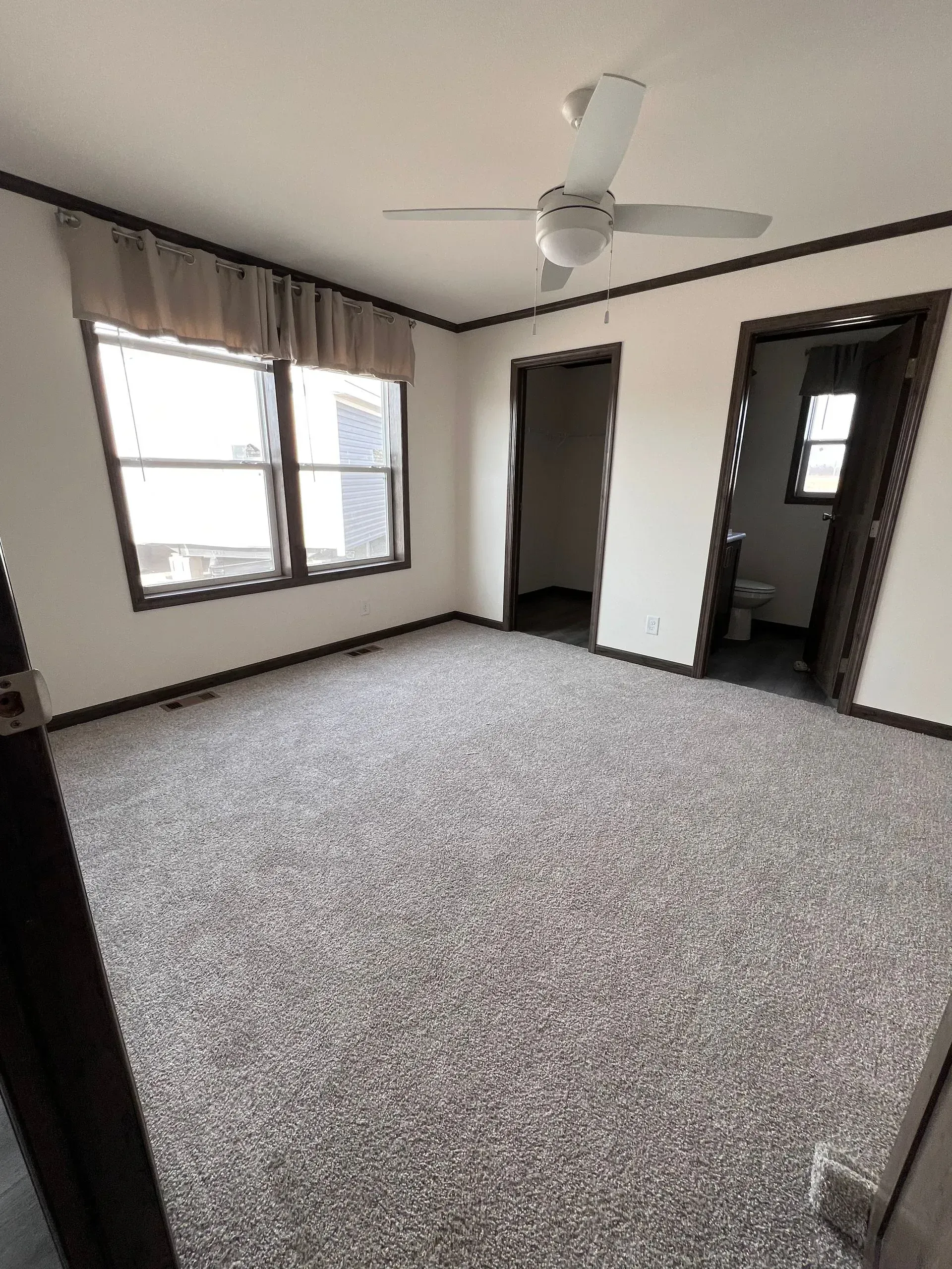 Empty room with light-colored carpet, window with brown trim and curtains, two doorways, and ceiling fan.