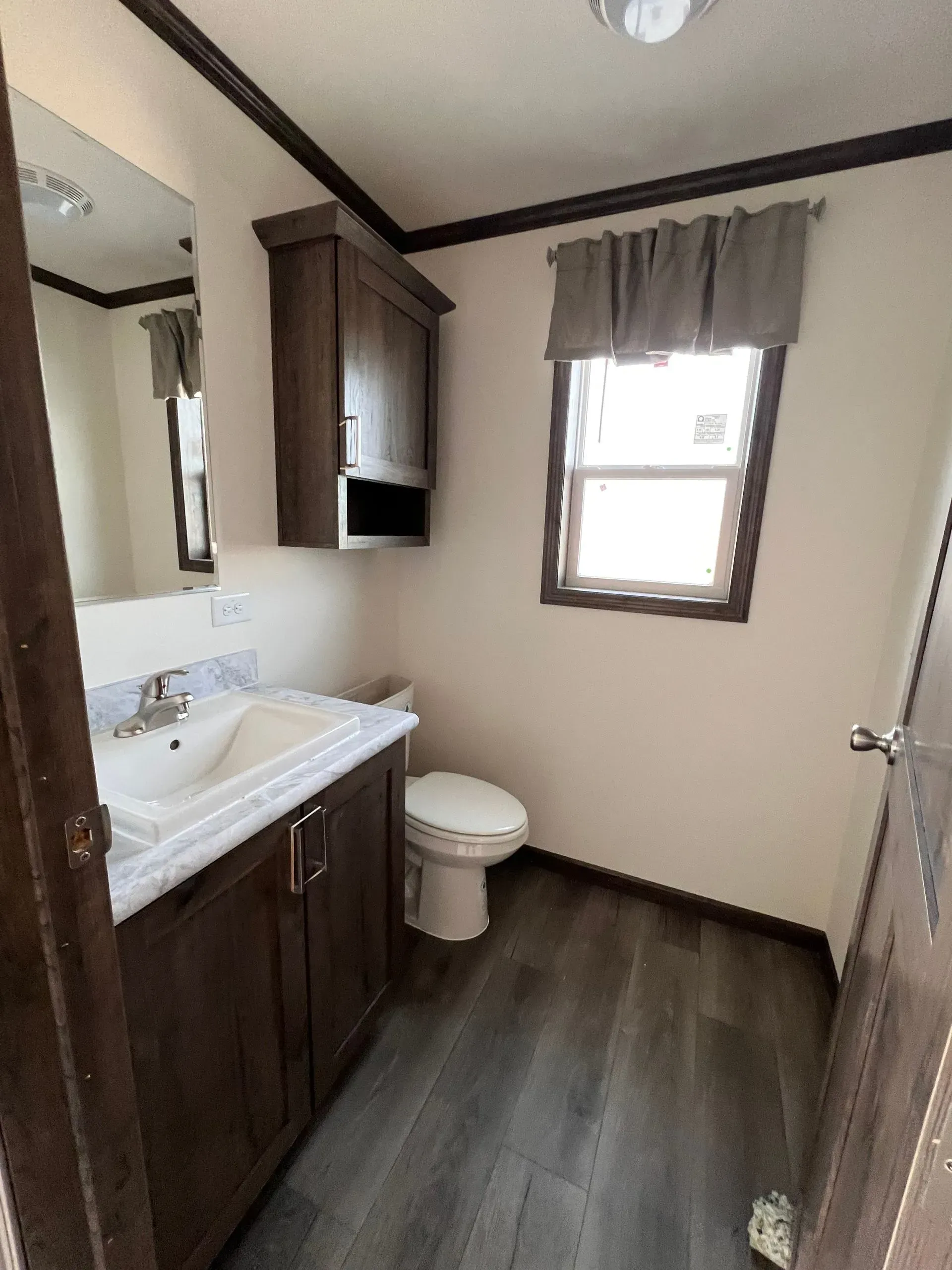 Bathroom with brown cabinetry, white sink and toilet, window with gray curtains, gray flooring.
