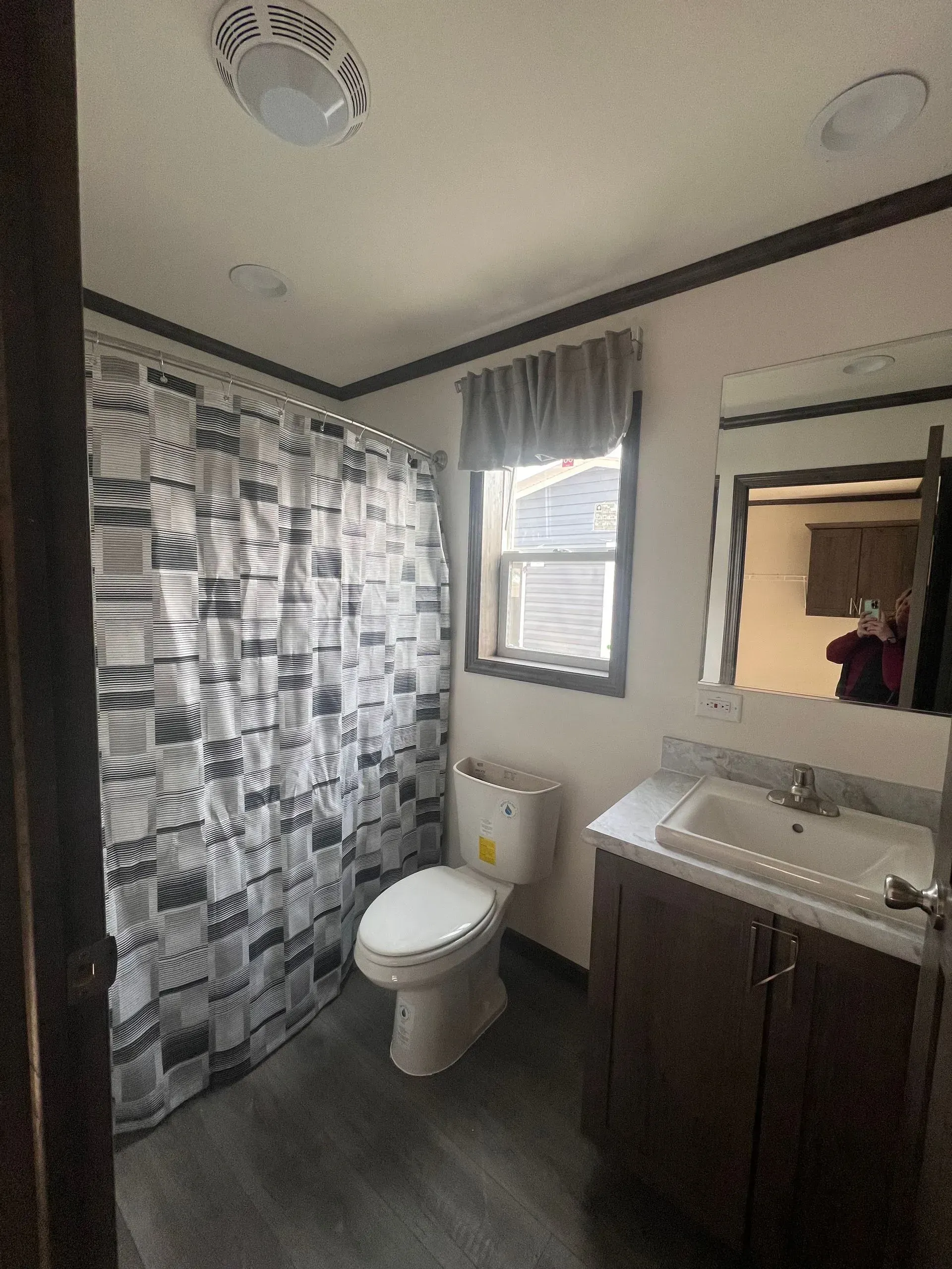 Bathroom with toilet, shower, vanity, window. Gray and white patterned shower curtain, dark brown trim.