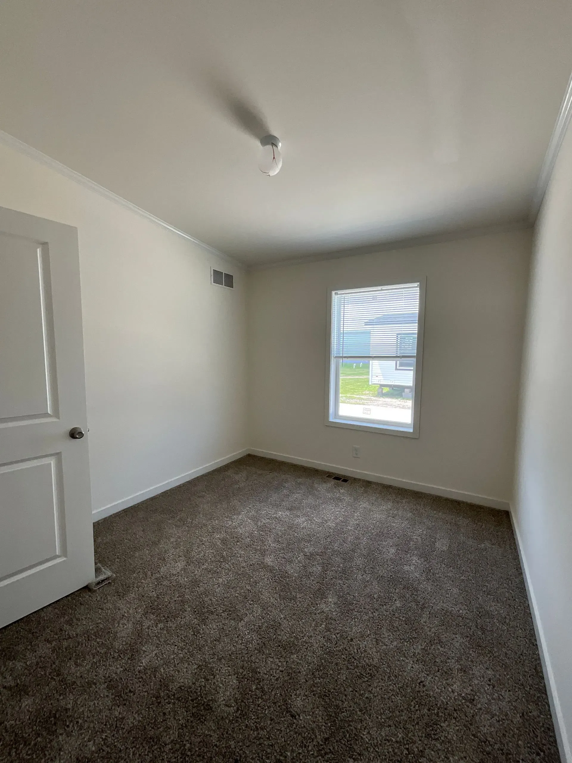 Empty room with white walls, brown carpet, window, and closed door.